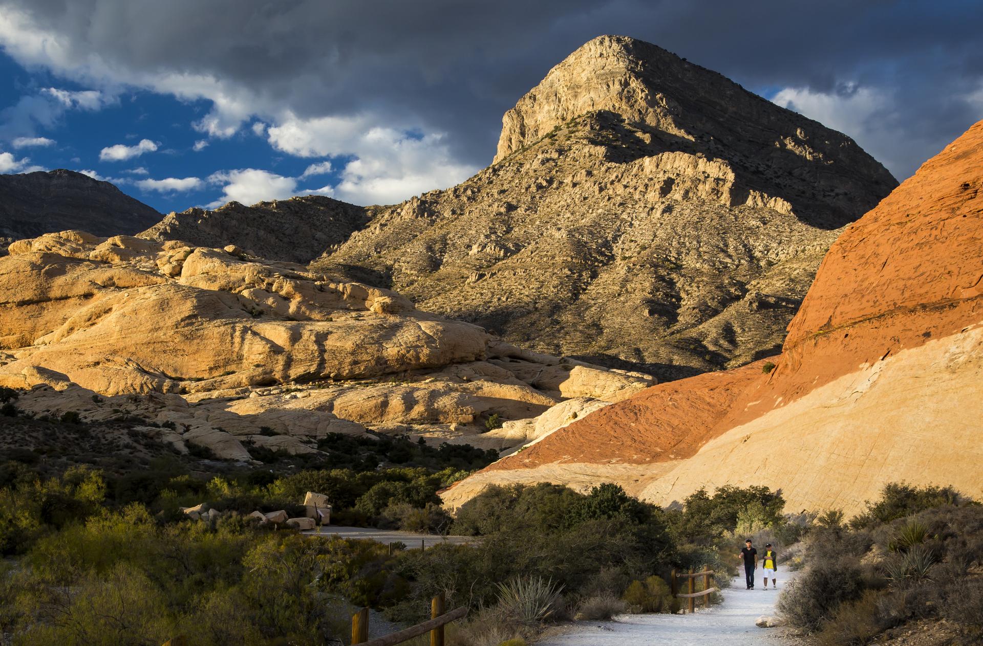 Two people walk on a path through dramatic, sunlit desert mountains under cloudy sky.