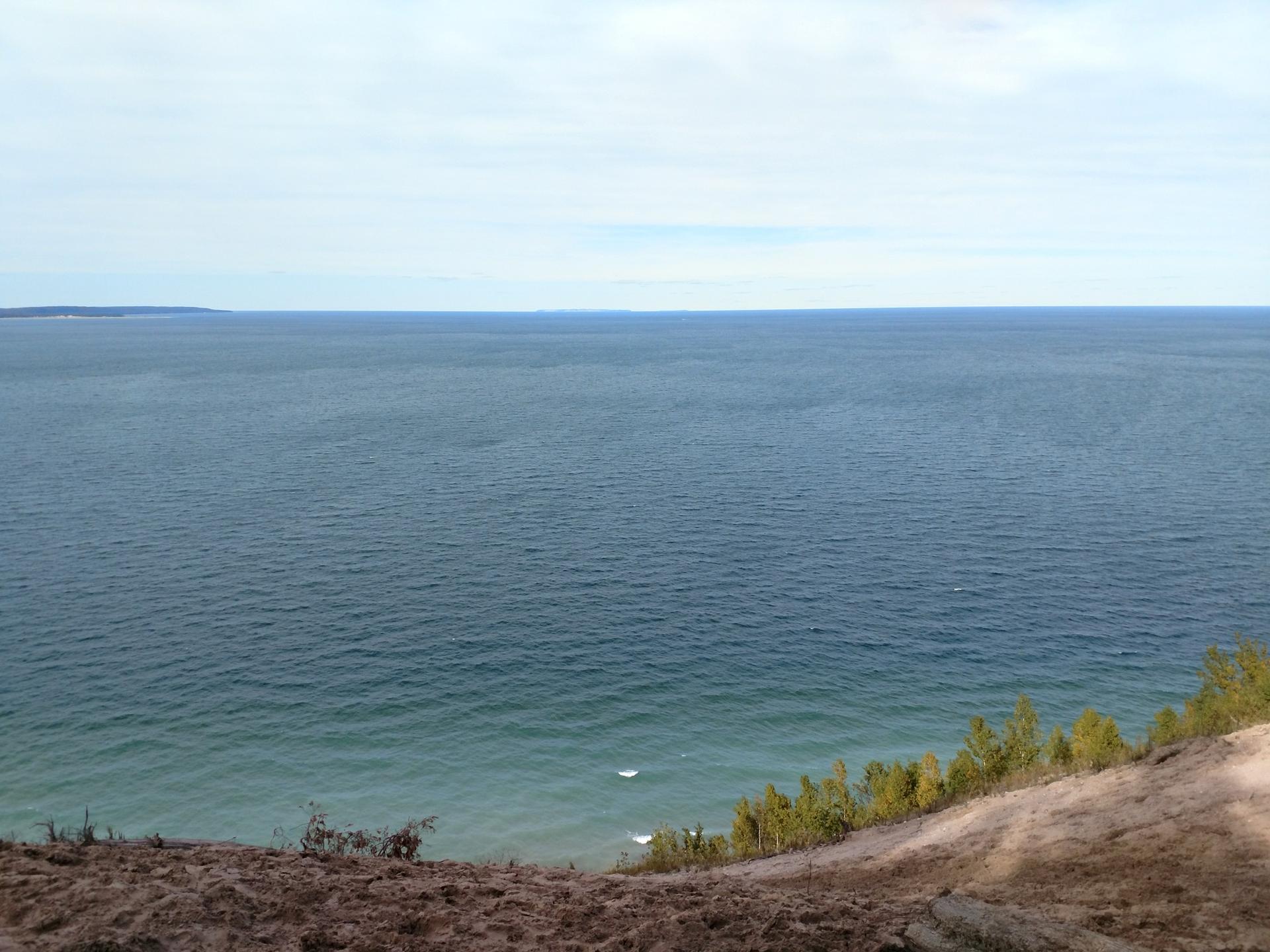 Expansive blue water from a sandy cliff with green trees under a bright sky.
