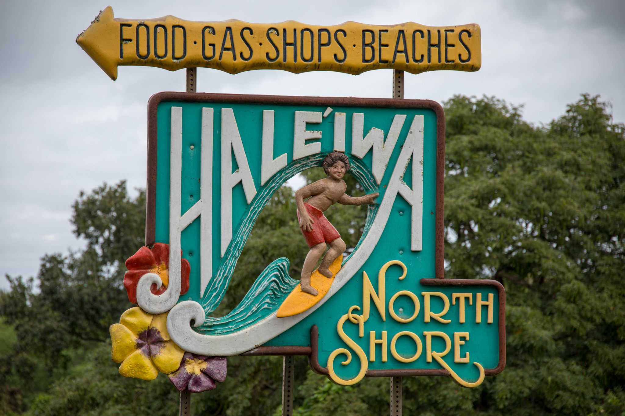 Colorful carved wooden sign featuring a surfer, set against green trees and cloudy sky. - Haleiwa Town – Haleiwa Town loop from Haleiwa