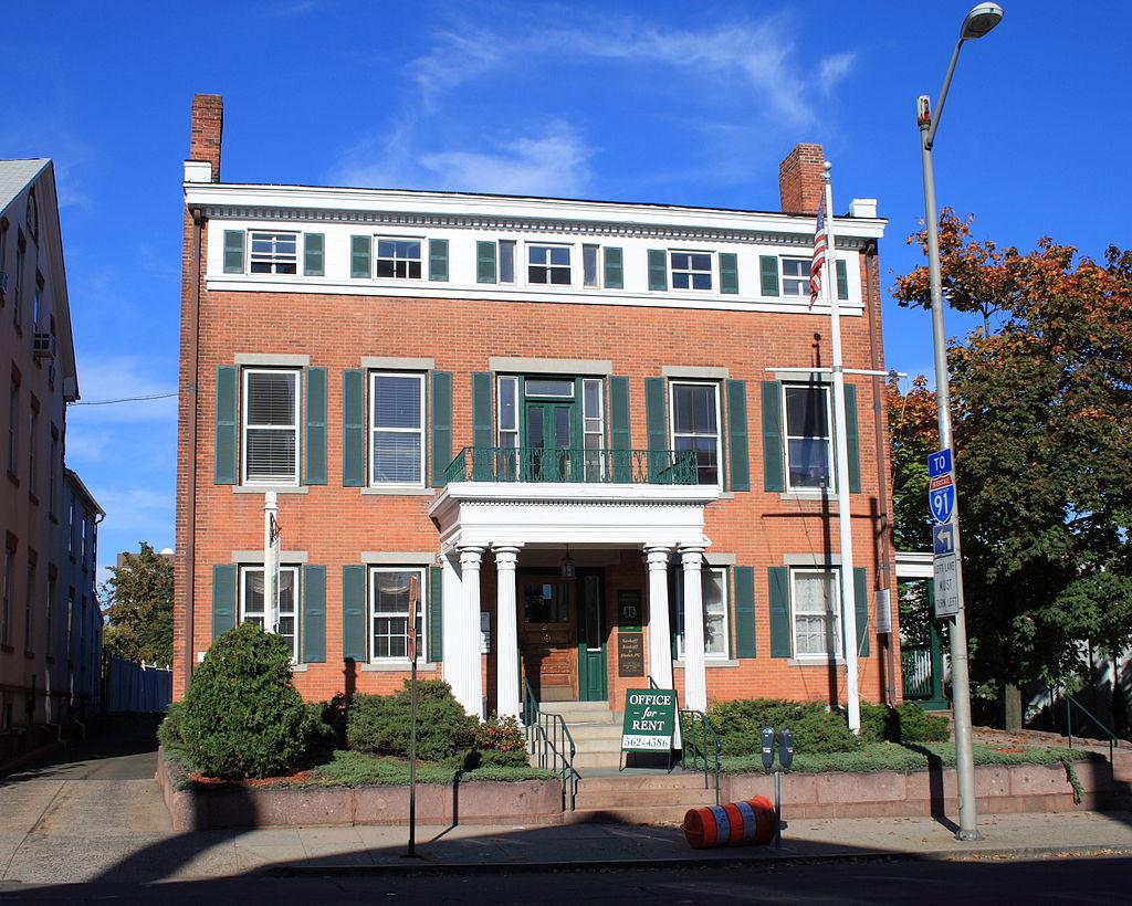 Historic red brick building with green shutters and a white porch on a sunny day.