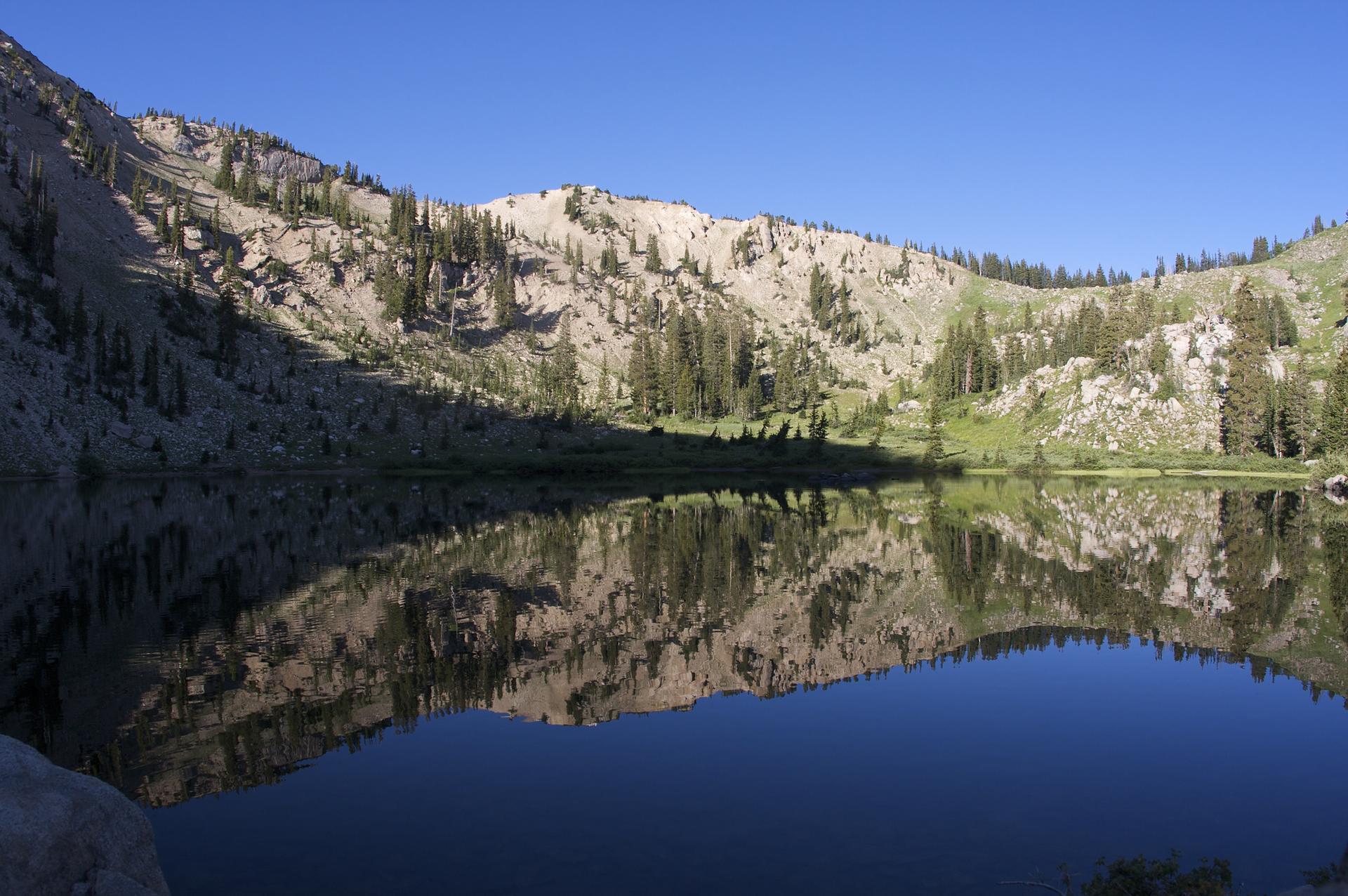 A still mountain lake perfectly reflecting steep, rocky mountains and pine trees.