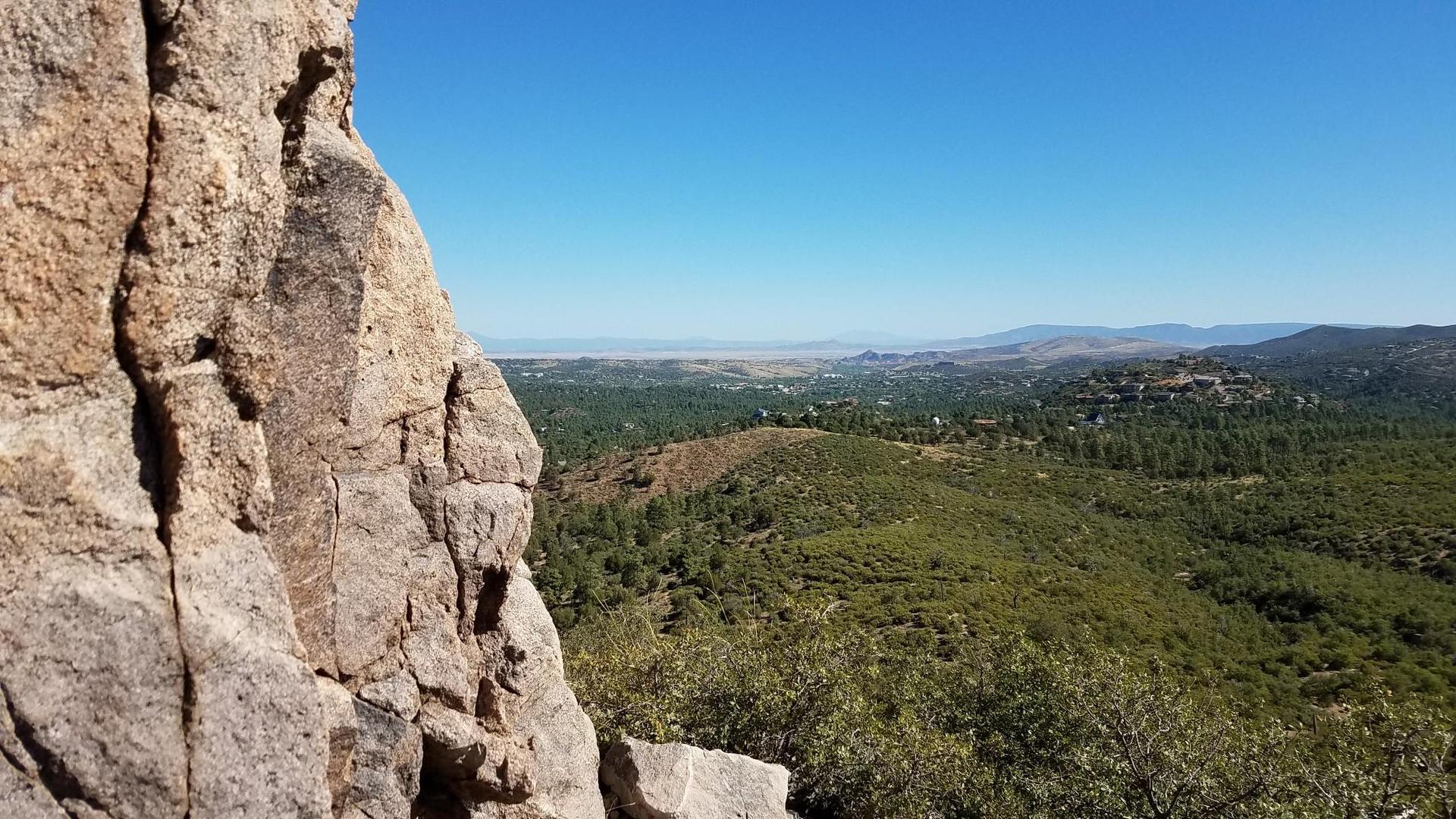 Large rock face overlooks a sunny, green forested valley and distant mountains.
