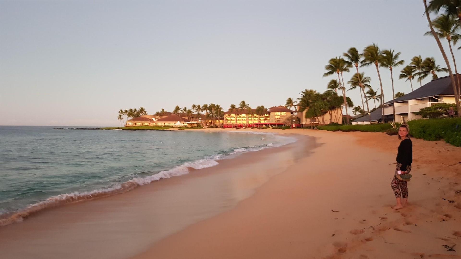 A person walks on a sandy beach at sunset, with resort buildings and palm trees.
