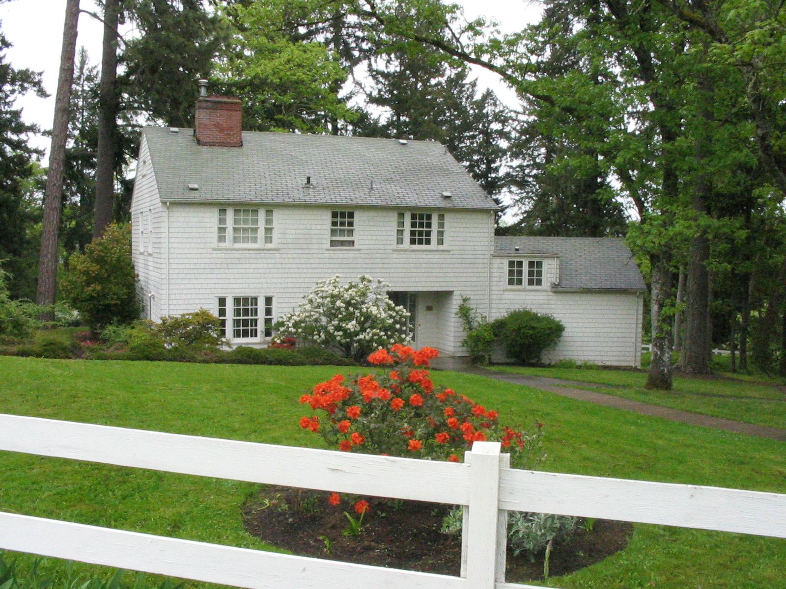 White house with green lawn, orange flowers, and white fence under overcast sky.