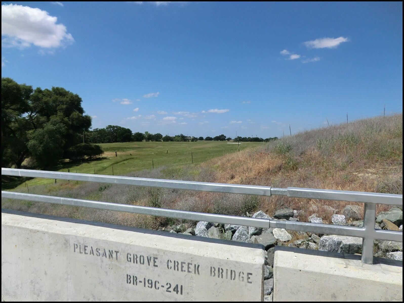 A bridge railing overlooks a sunny green field with distant trees under a blue sky.