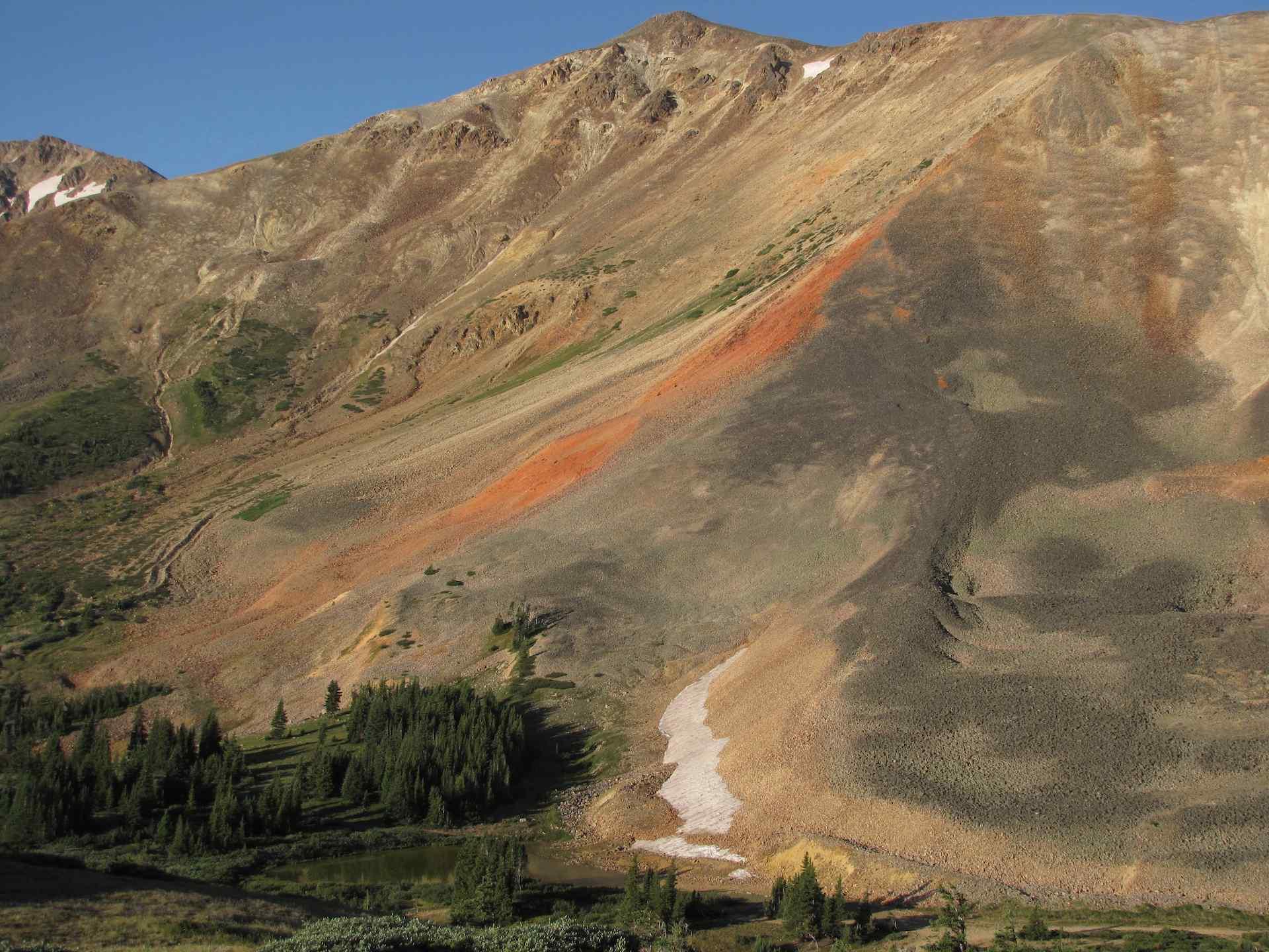 Sunny mountain landscape showing colorful rock, green vegetation, and a patch of snow.