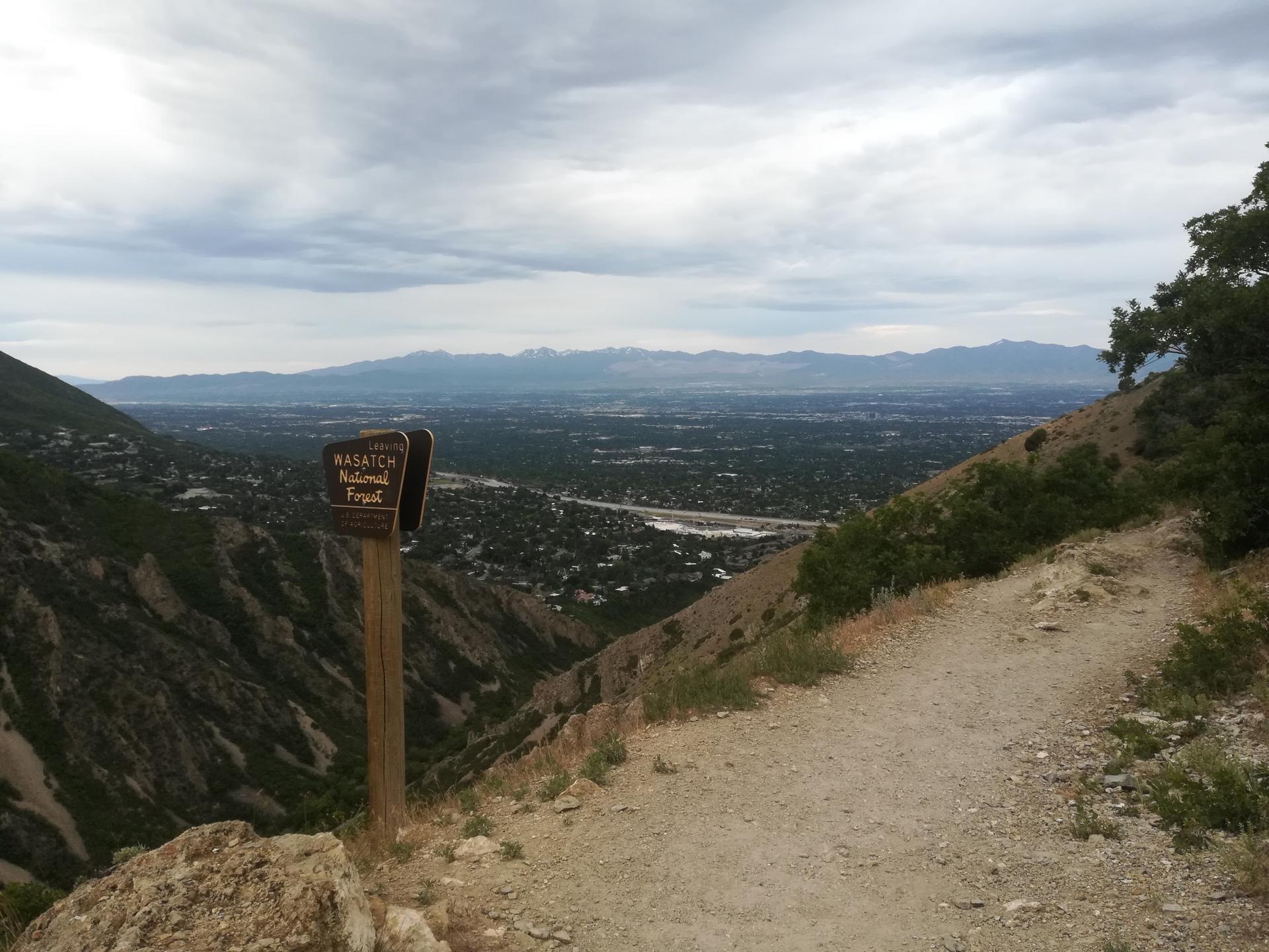 Dirt path and sign overlooking a valley town and mountains under cloudy skies.