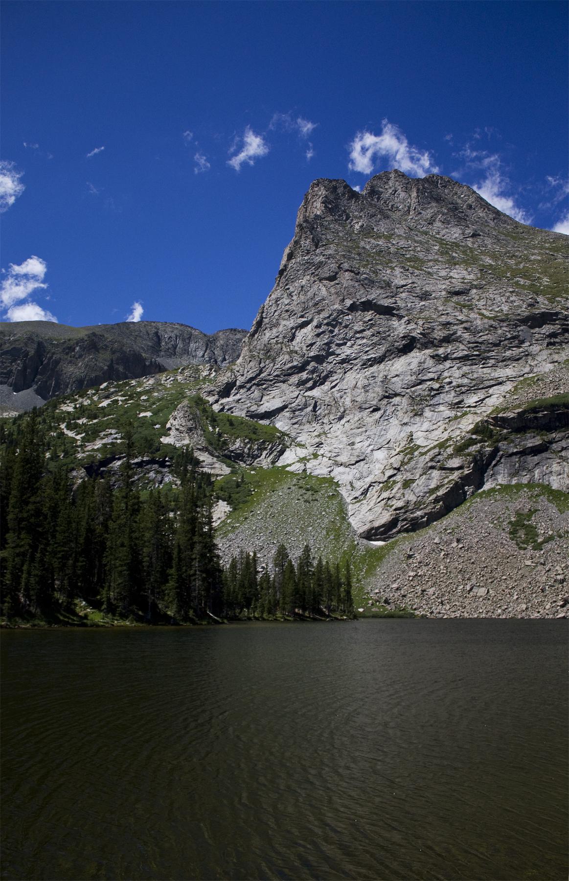 Dark lake in foreground, large rocky mountain under a bright blue sky.