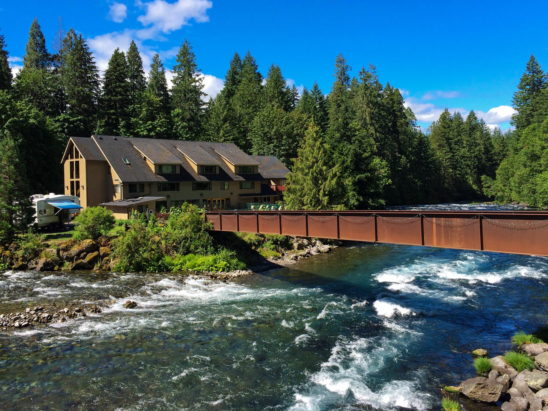 Large building and metal bridge over a rushing river, surrounded by forest.