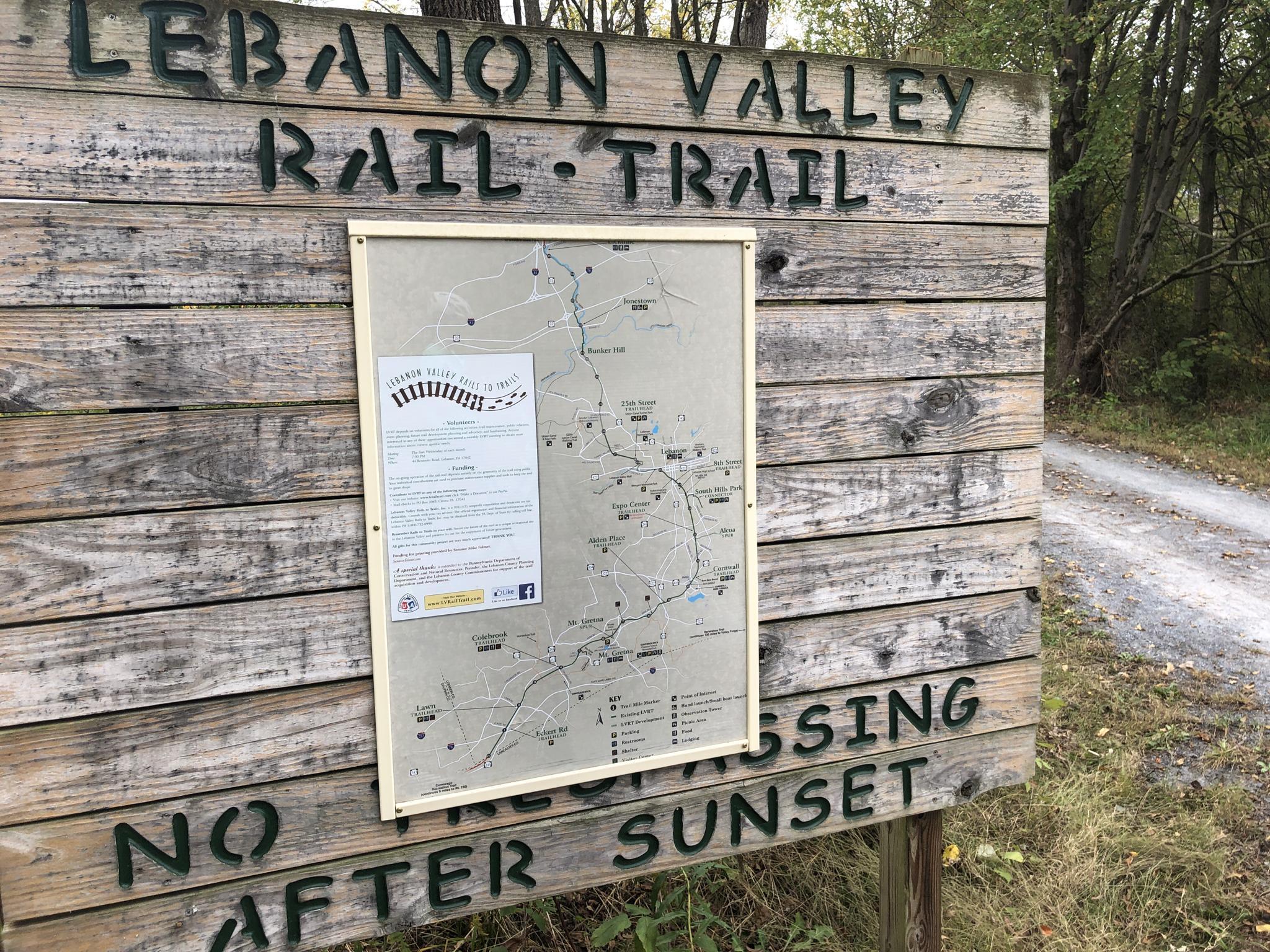 A wooden trail sign with a map stands next to a dirt path.