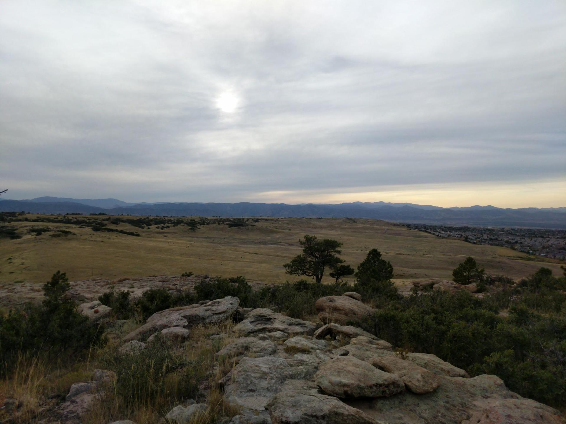Rocky foreground overlooks golden fields and distant mountains under an overcast sky.