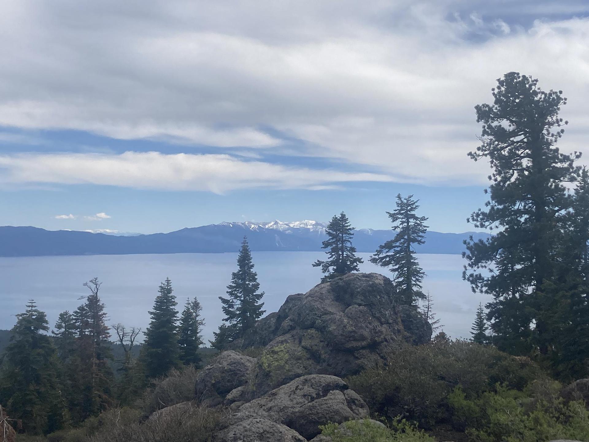 A large lake with distant snow-capped mountains, framed by rocks and pine trees.