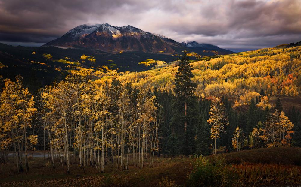 Sunlit golden autumn forests below a dramatic, snow-capped mountain under dark clouds.