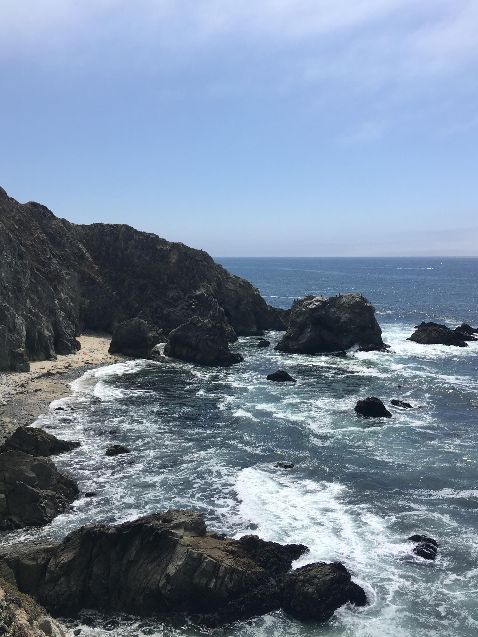 Coastal cliffs, rocky beach, and ocean waves under a bright sky.