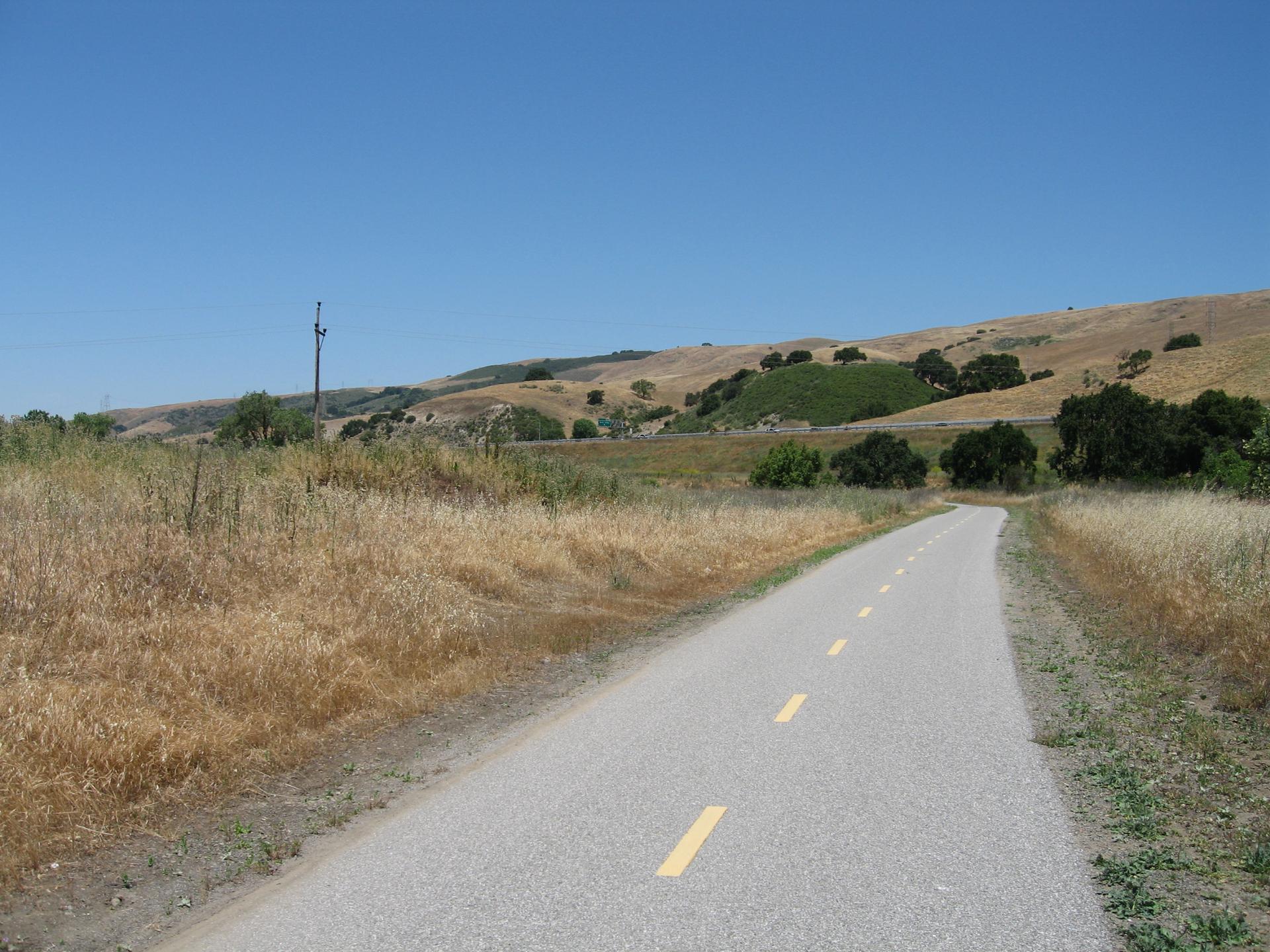 Paved path through dry golden hills under a clear blue sky.