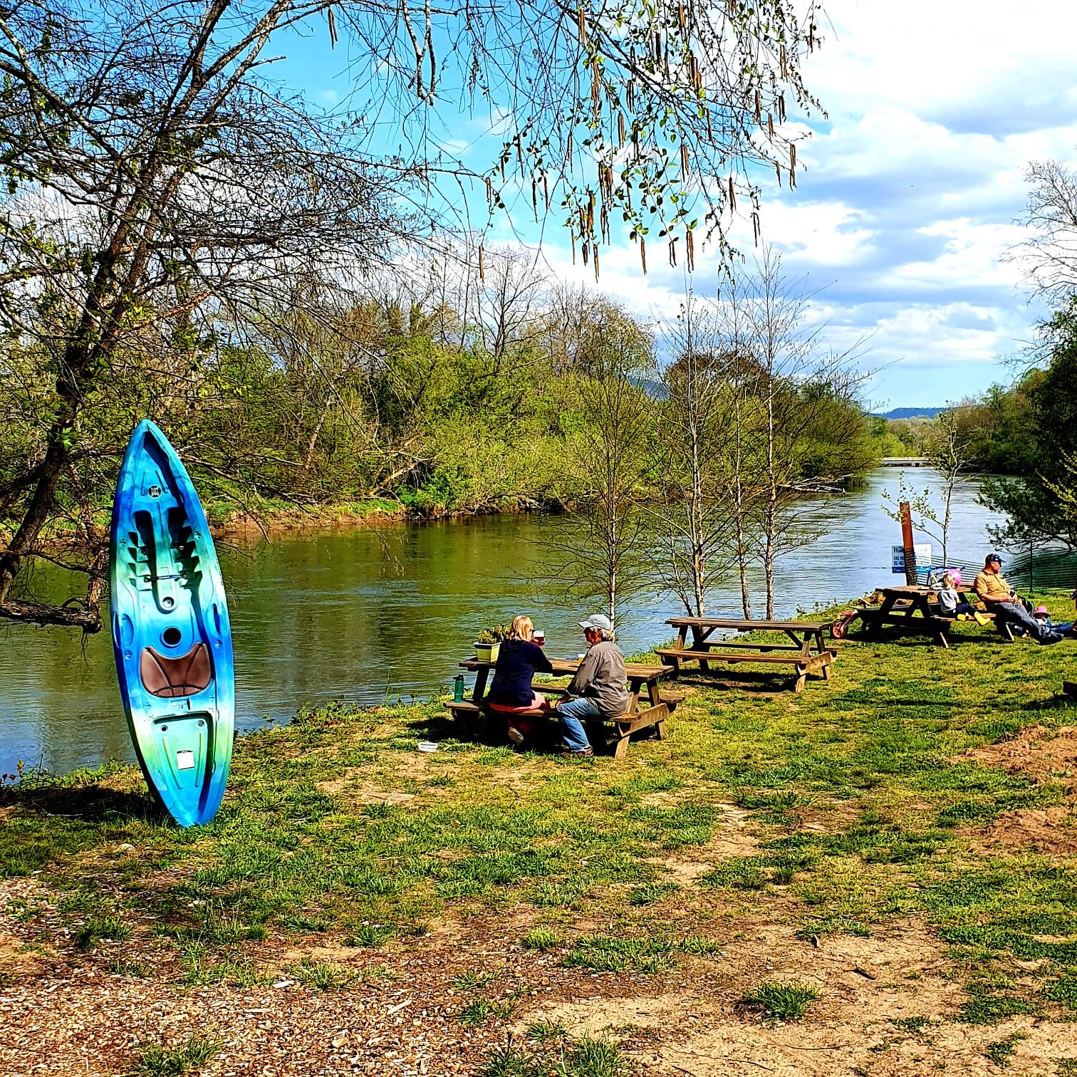 People relaxing at picnic tables by a river with a colorful kayak.