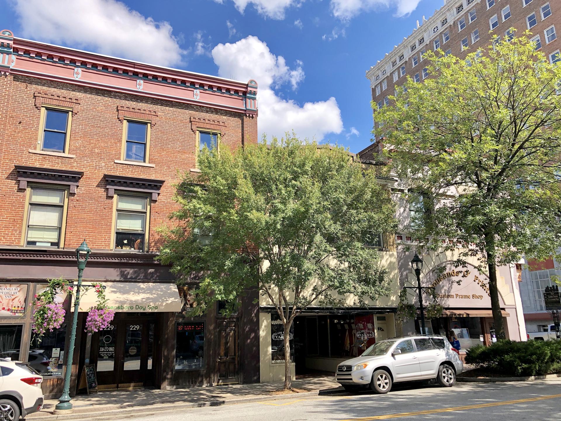 Urban street with brick buildings, parked cars, and green trees under bright sky.