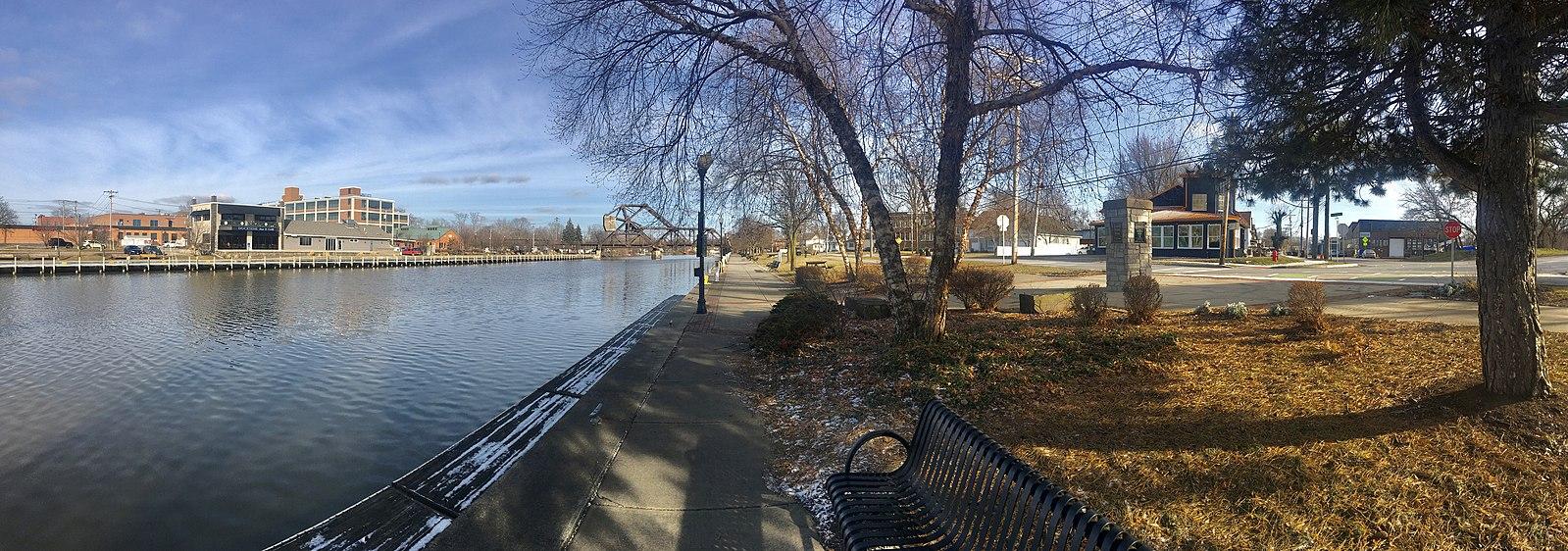 Waterfront with buildings, bare trees, and a bench on a sunny day.