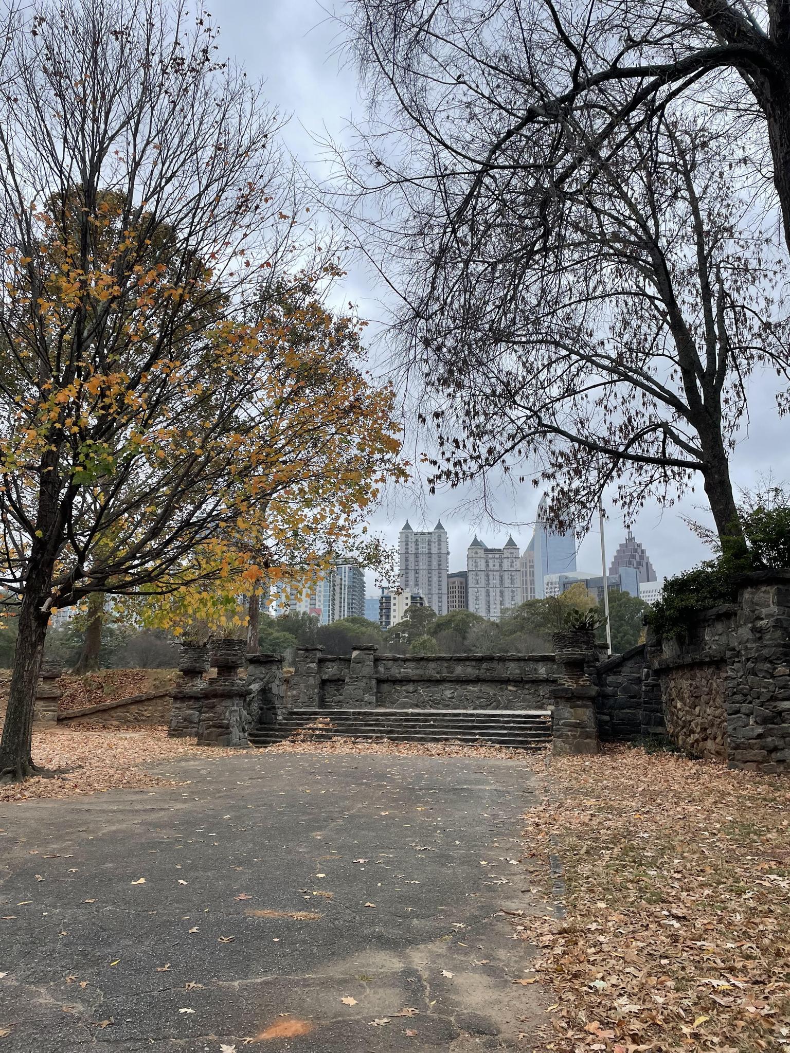 Stone structure and autumn trees with a city skyline under a gray sky.