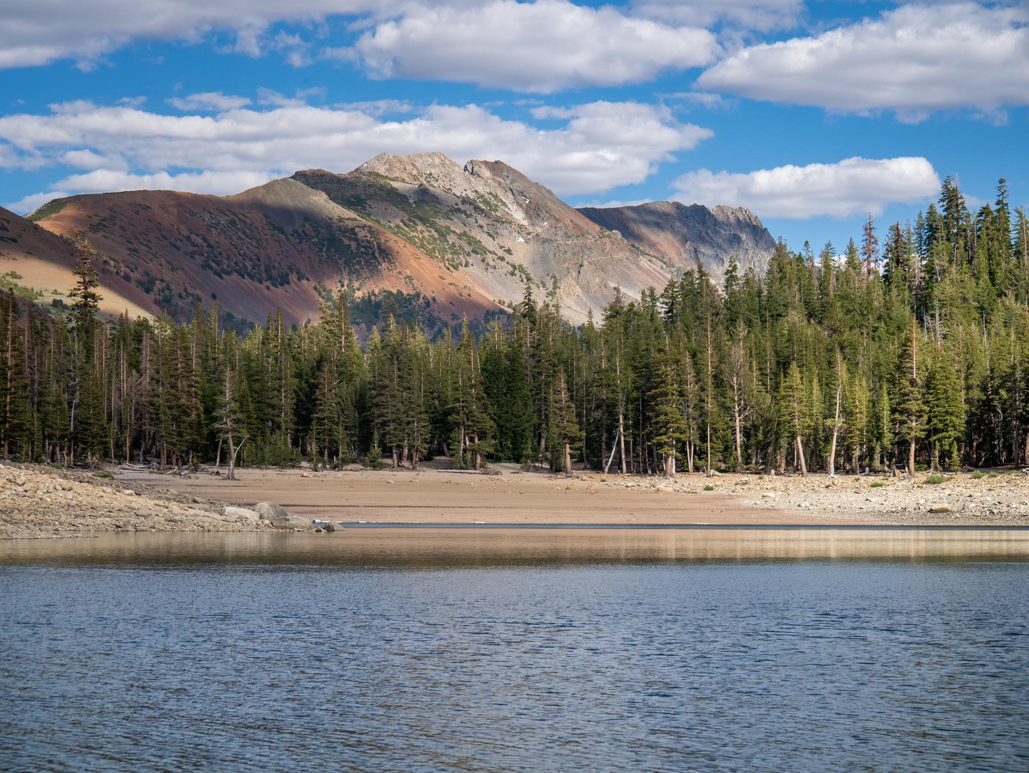 Blue lake, dry shore, pine forest, and mountains under a partly cloudy sky.