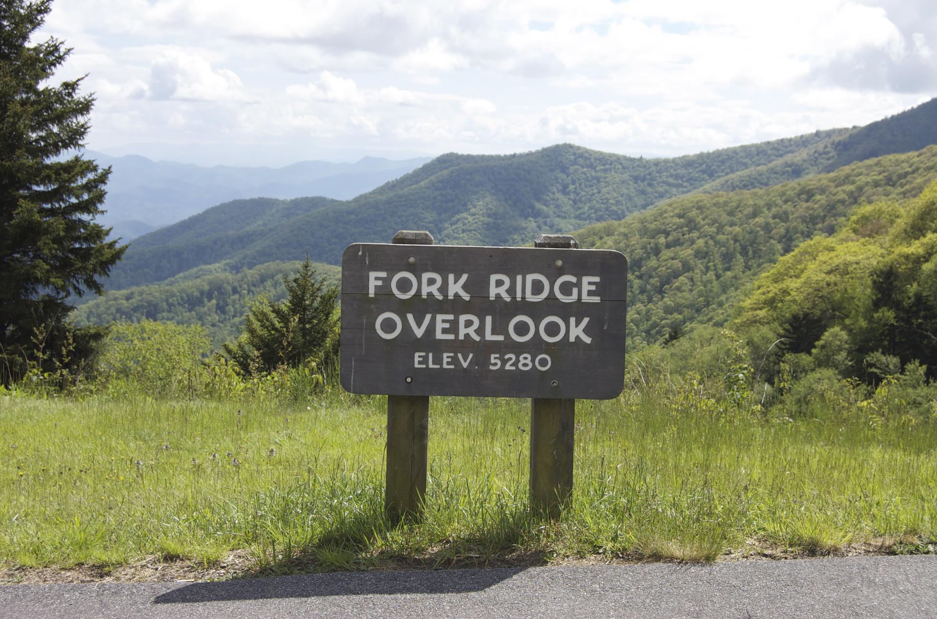 A weathered wooden sign with white text, overlooking rolling green mountains.