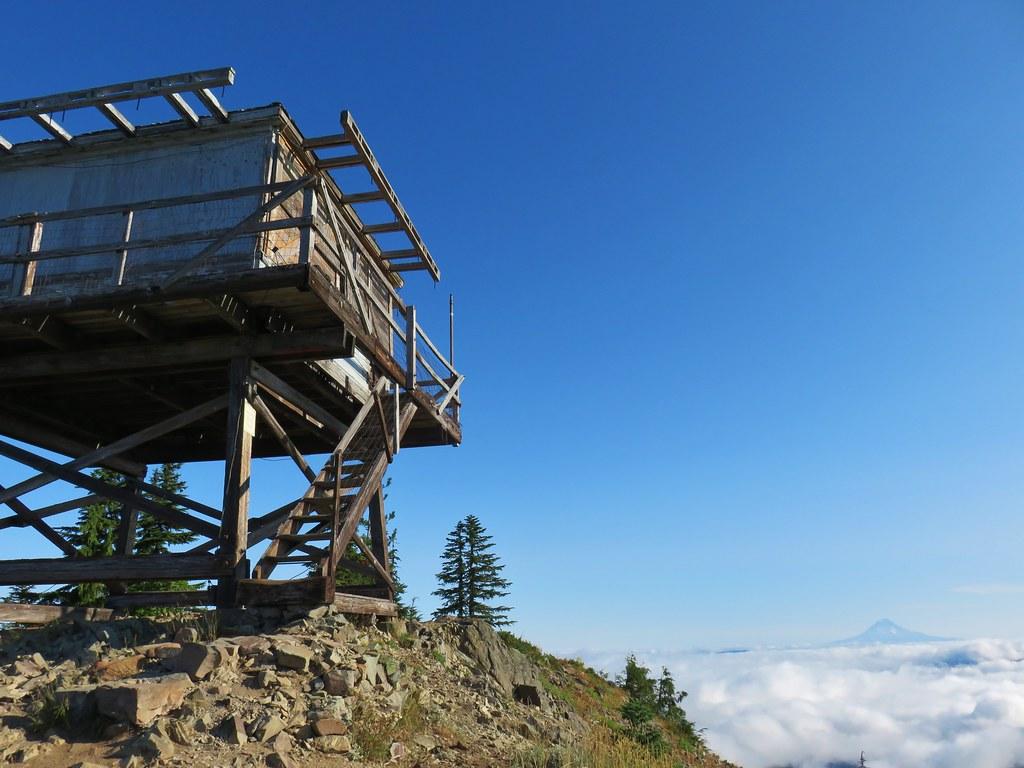 A wooden fire lookout stands on a sunny mountain peak above a sea of clouds.