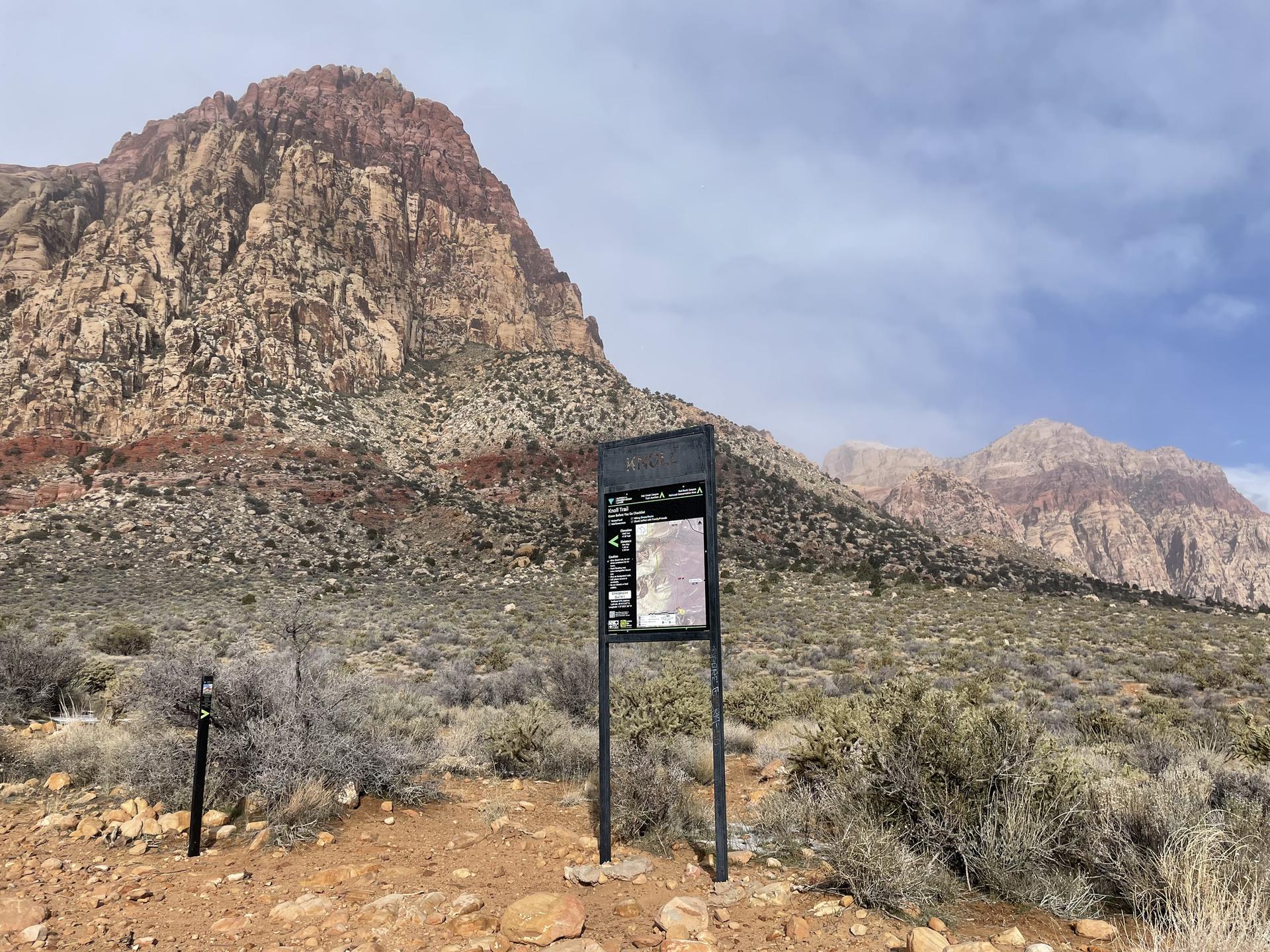 Trailhead sign in desert landscape with a massive red rock mountain under cloudy sky.