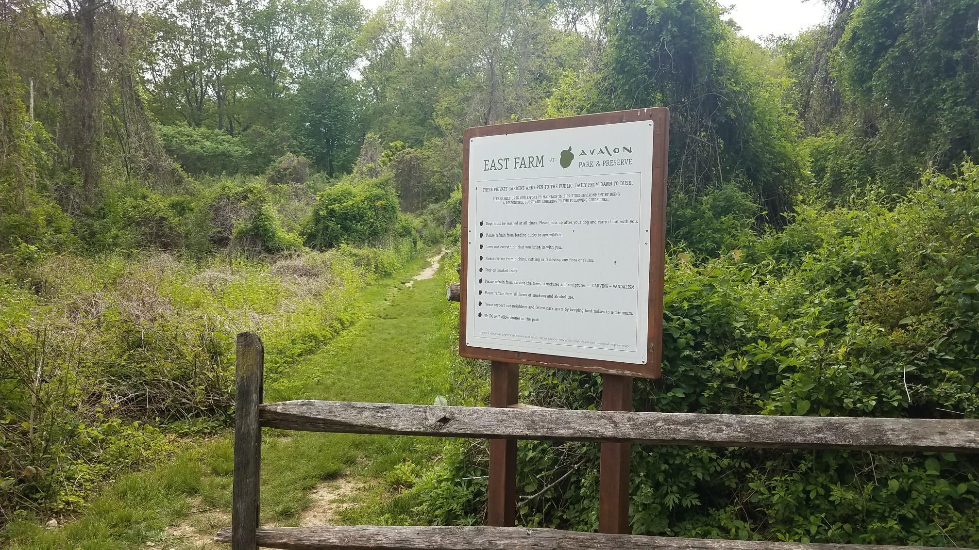 A white sign and wooden fence beside a grassy path in a sunny, overgrown landscape.