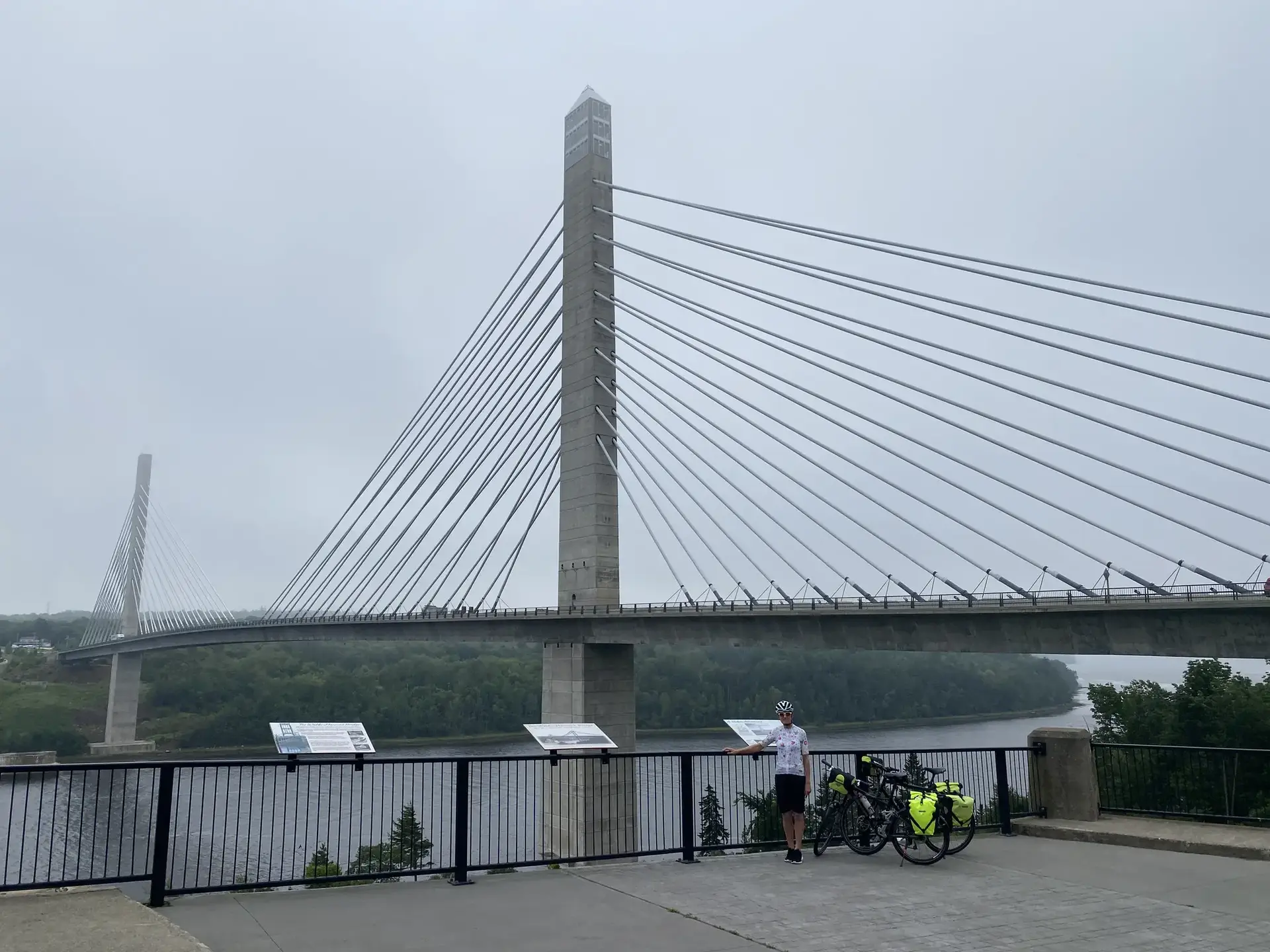 Cyclist with bicycle standing by a large cable-stayed bridge on an overcast day.