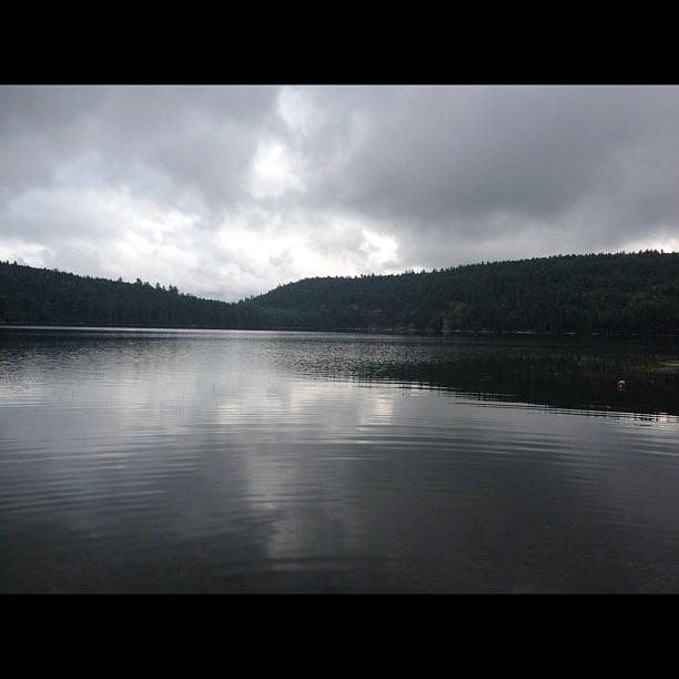 Dark, calm lake reflecting overcast sky, surrounded by forested hills.