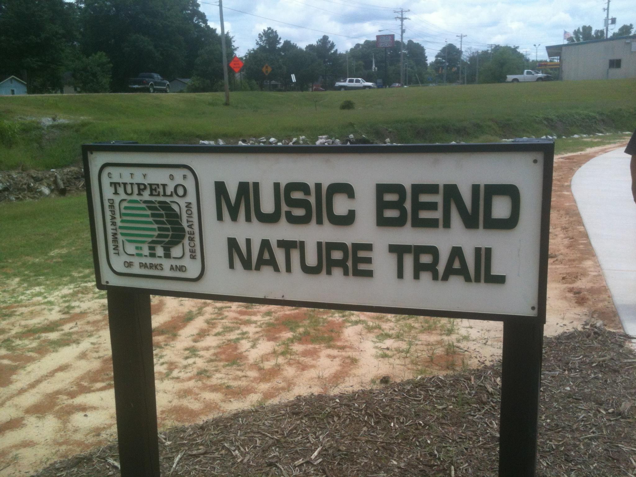 A white sign with dark lettering stands beside a paved path and grassy area.