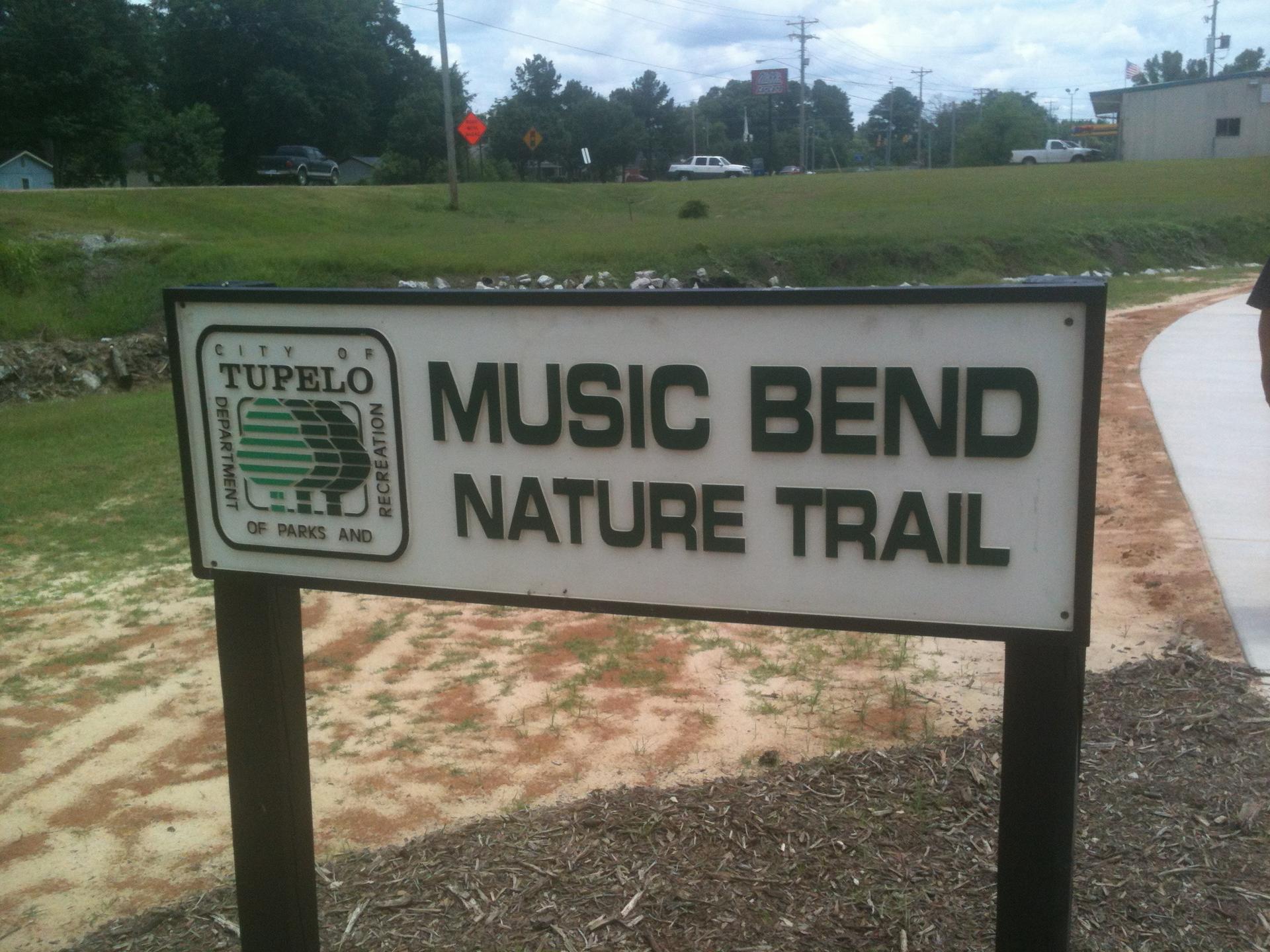 A white sign with dark lettering stands beside a paved path and grassy area.