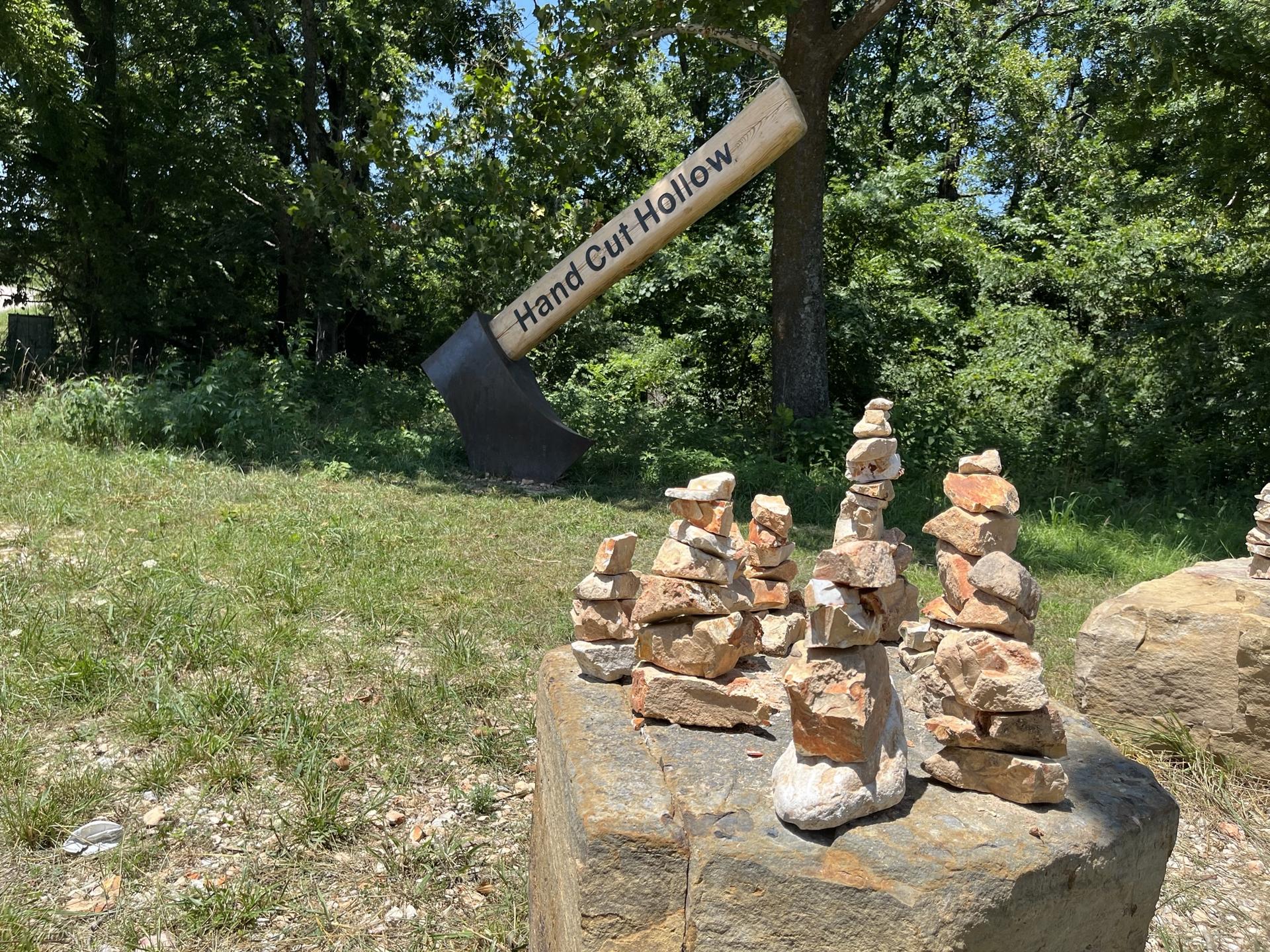 Rock cairns sit on a boulder with a large axe sculpture behind.