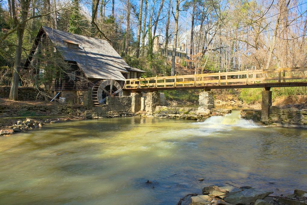 A rustic wooden mill with a water wheel next to a flowing river and bridge.