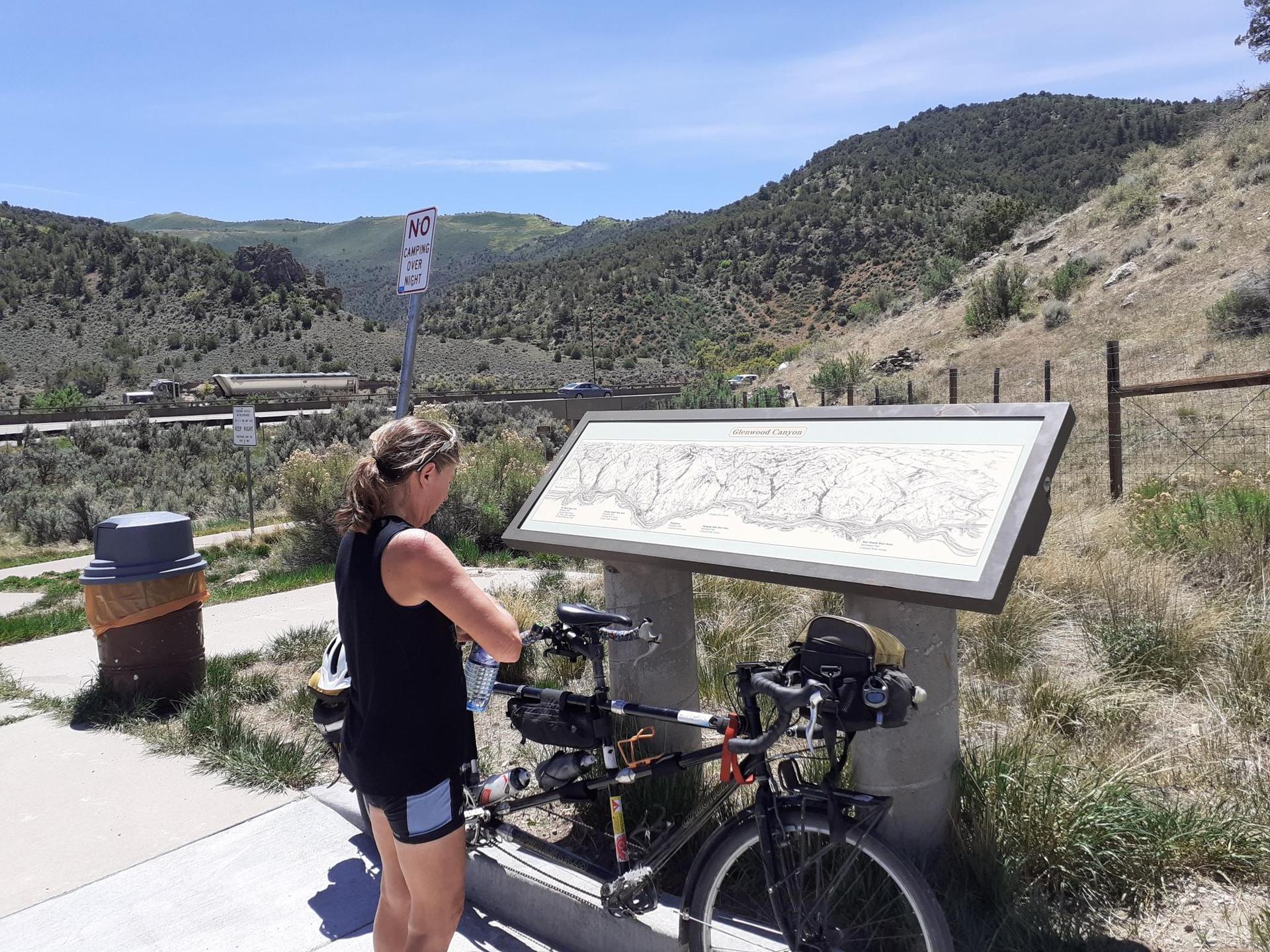 Woman standing by a tandem bicycle, looking at an informational sign on a sunny day.
