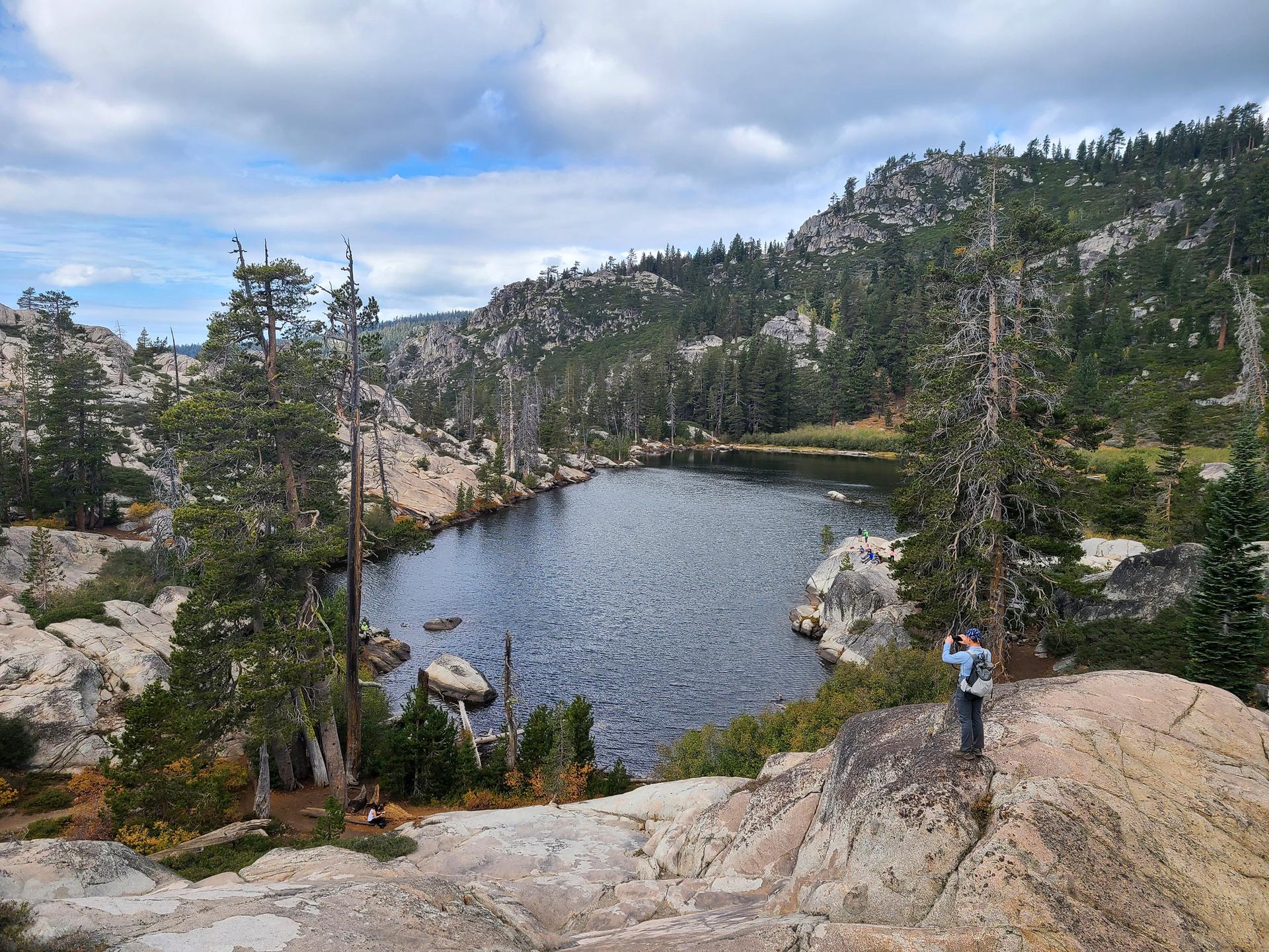 Person on rocky cliff overlooking a lake surrounded by pine trees under cloudy sky.