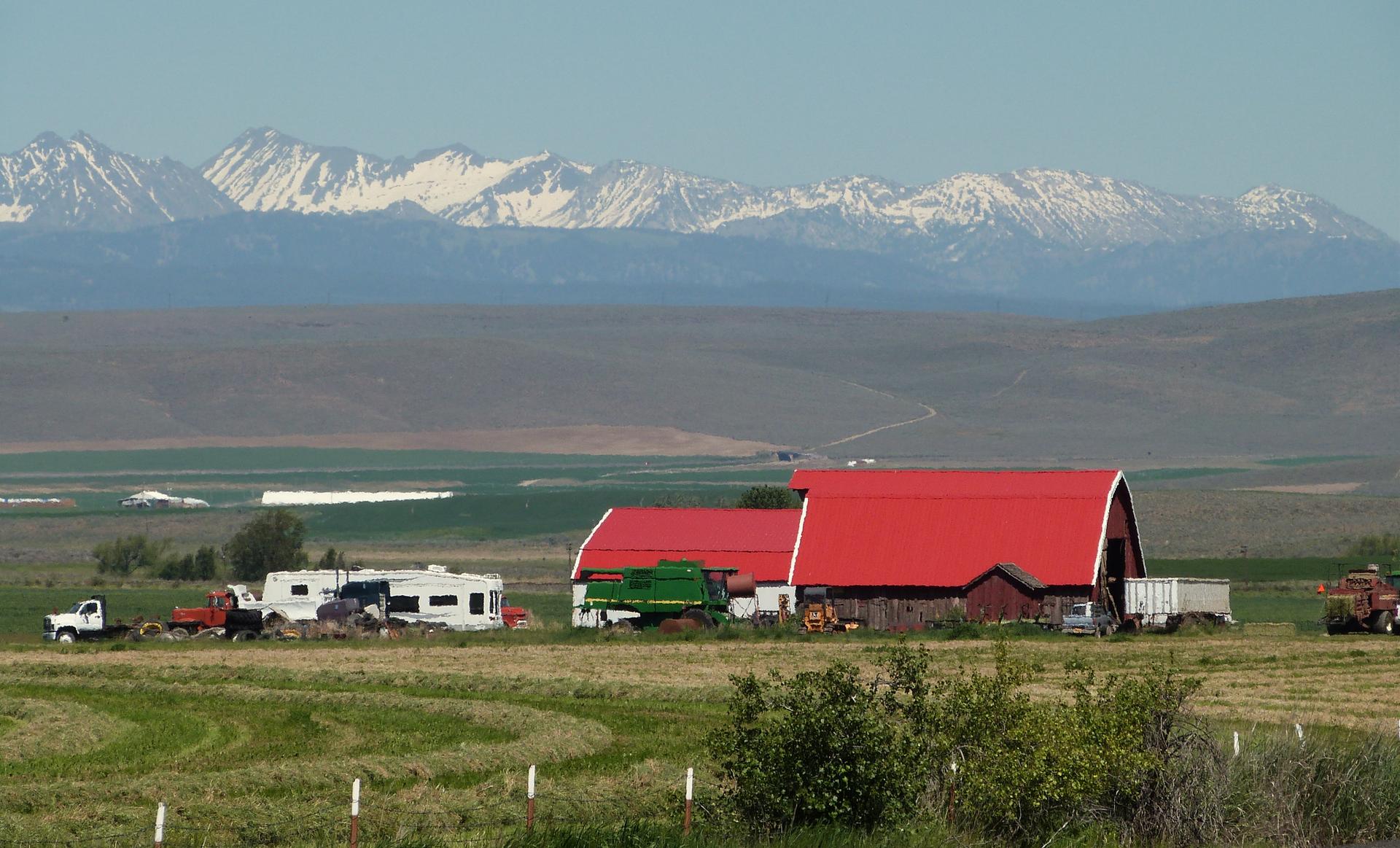 Red barn and farm equipment in a green field with snow-capped mountains.