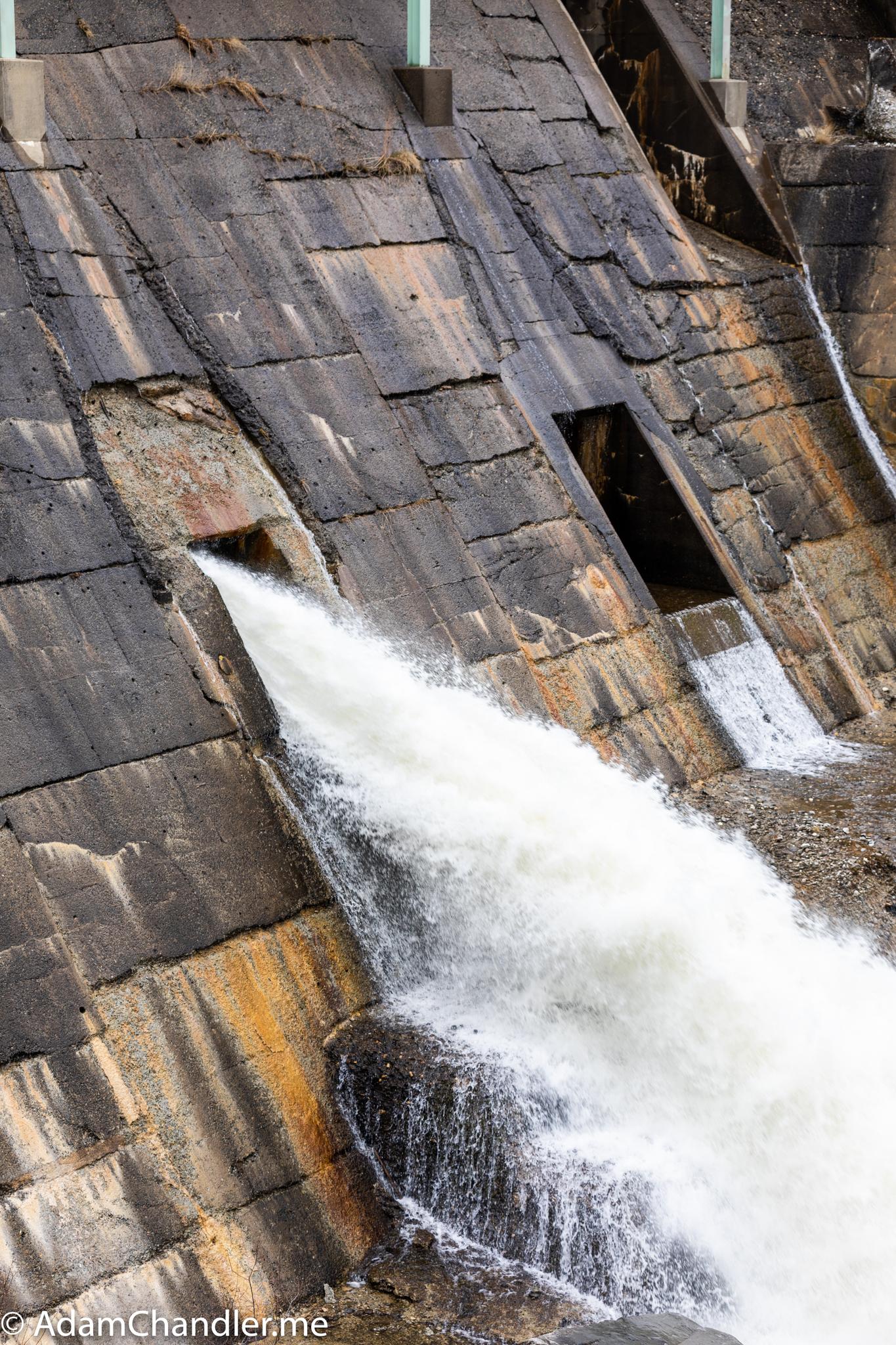 Water gushes from an opening in a large, textured concrete dam.