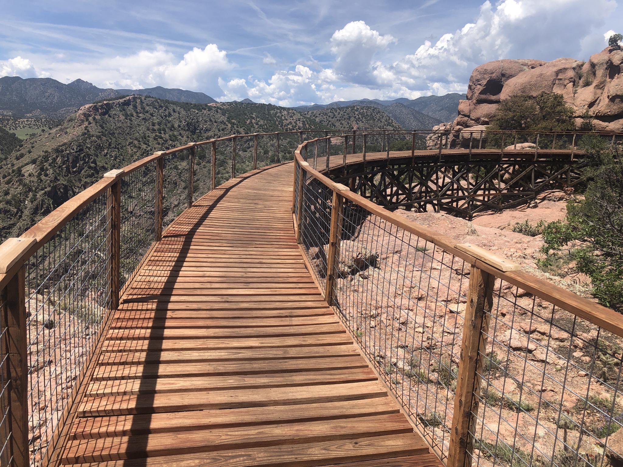 Curved wooden walkway with mesh fencing overlooking a wooden trestle and arid hills.