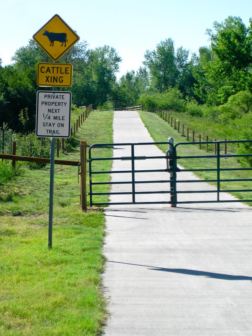 Paved trail with a closed gate, warning signs, and green trees under bright sun.