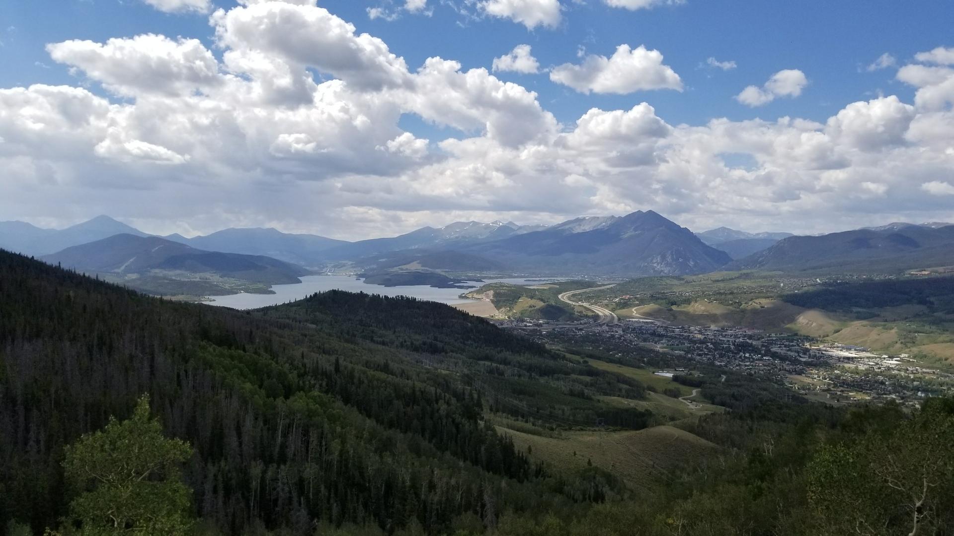 Panoramic view of a mountain lake, forested hills, and a town.