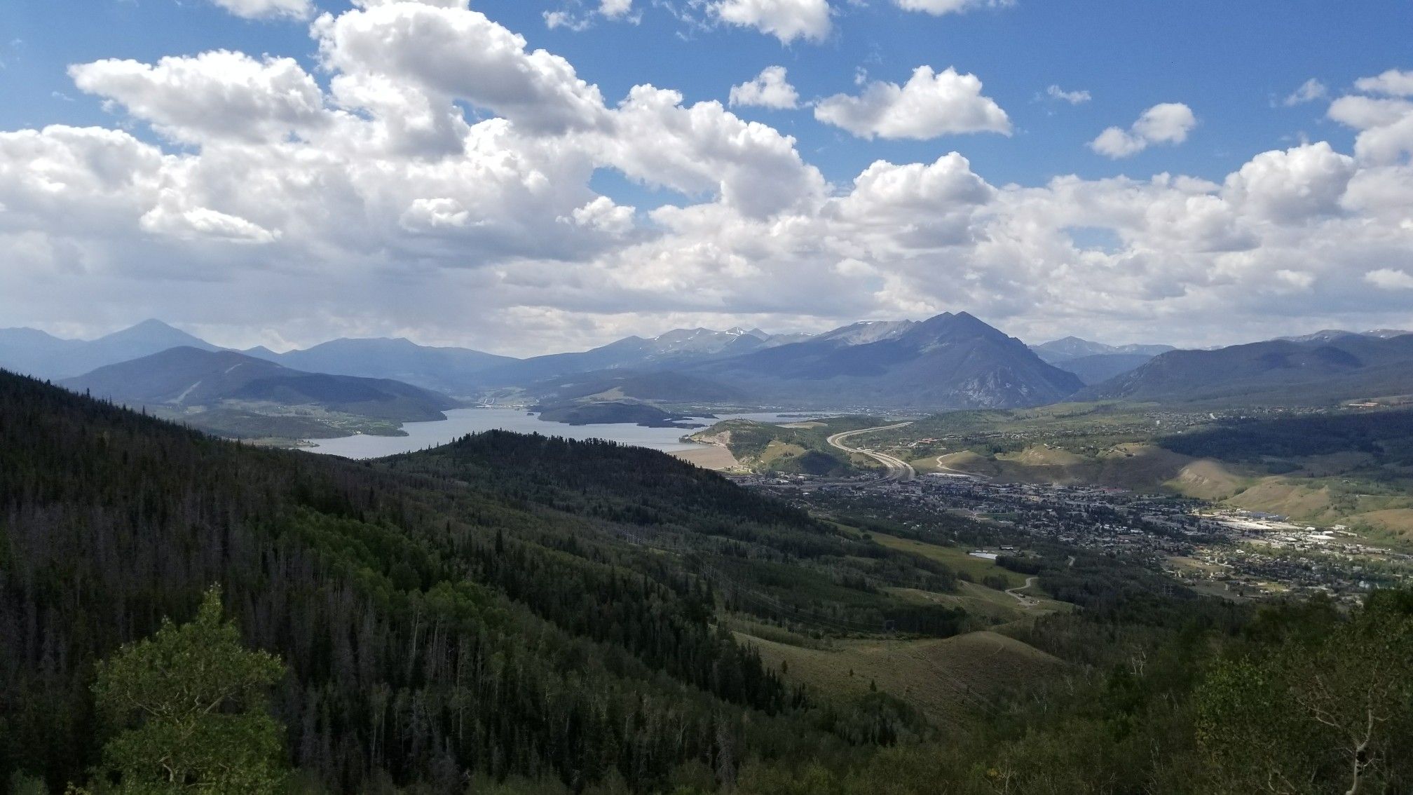Panoramic view of a mountain lake, forested hills, and a town. - Ptarmigan Loop Trail