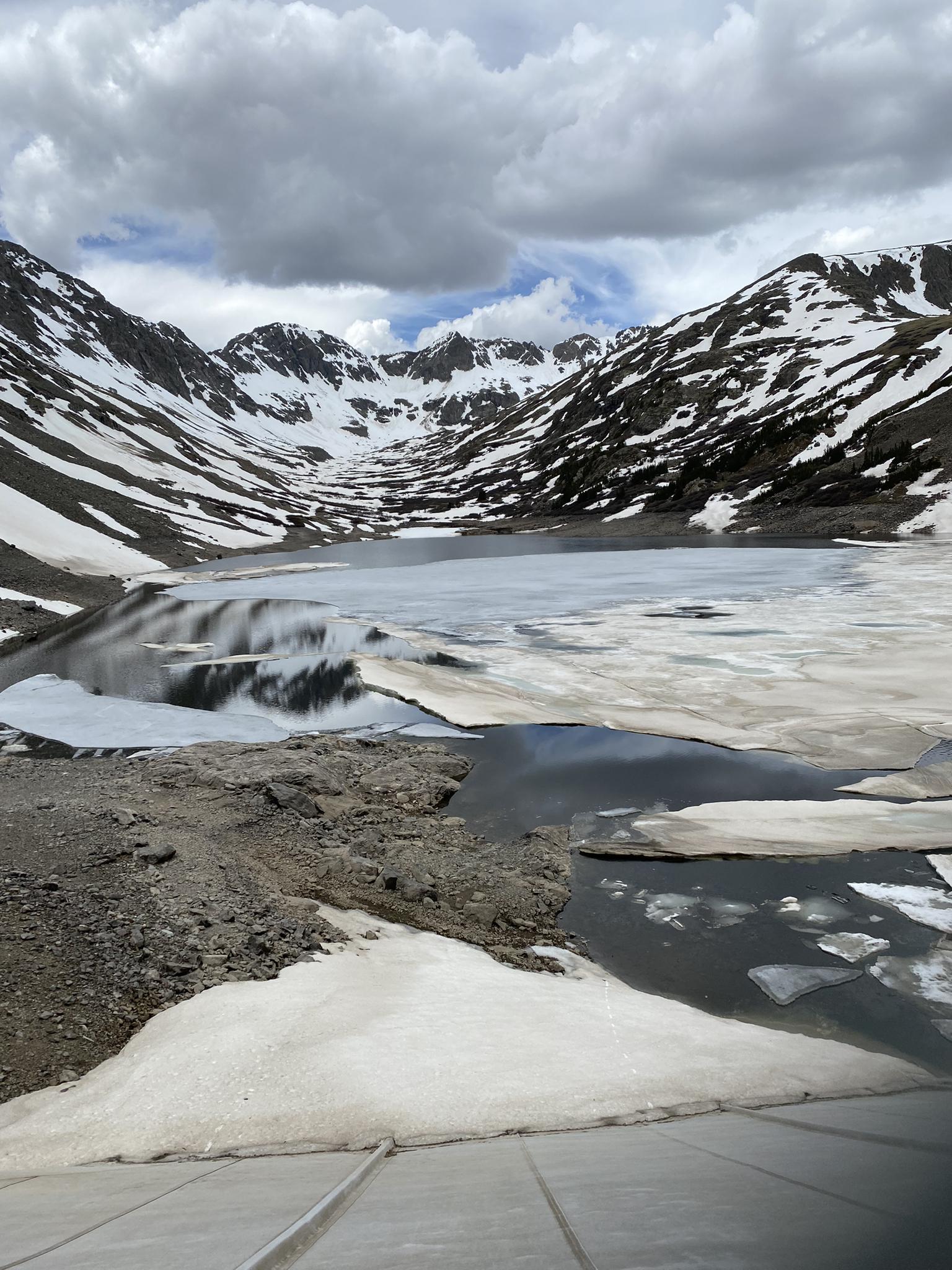 Partially frozen mountain lake surrounded by snow-dusted peaks under a cloudy sky.