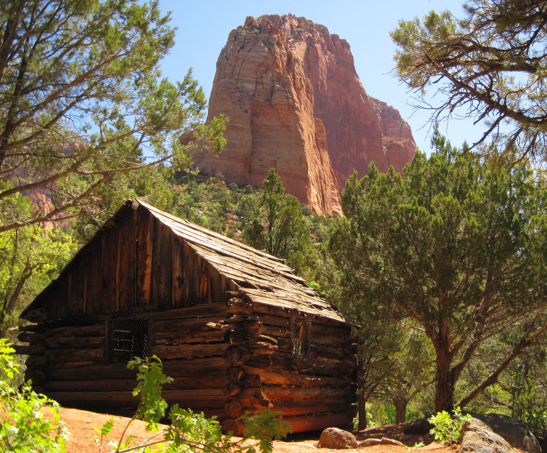 An old wooden cabin sits among green trees with a large red mountain behind.