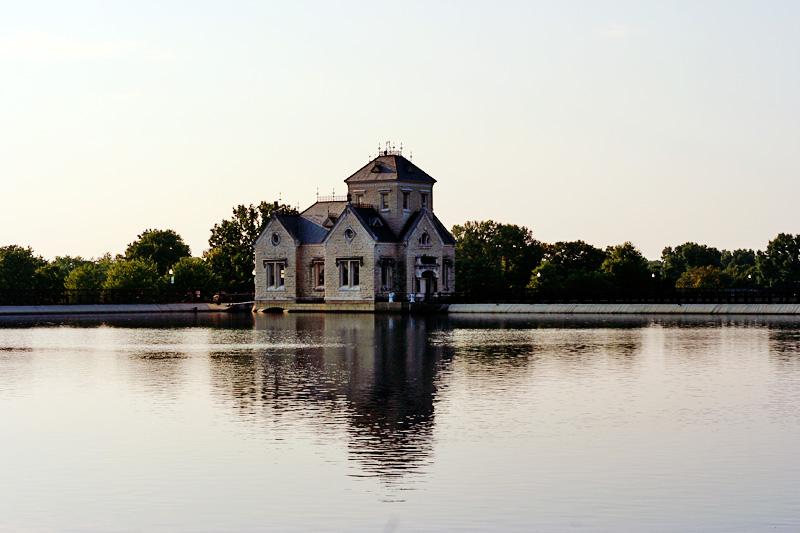 Stone building with a tower stands in calm water, trees in background, bright sky.