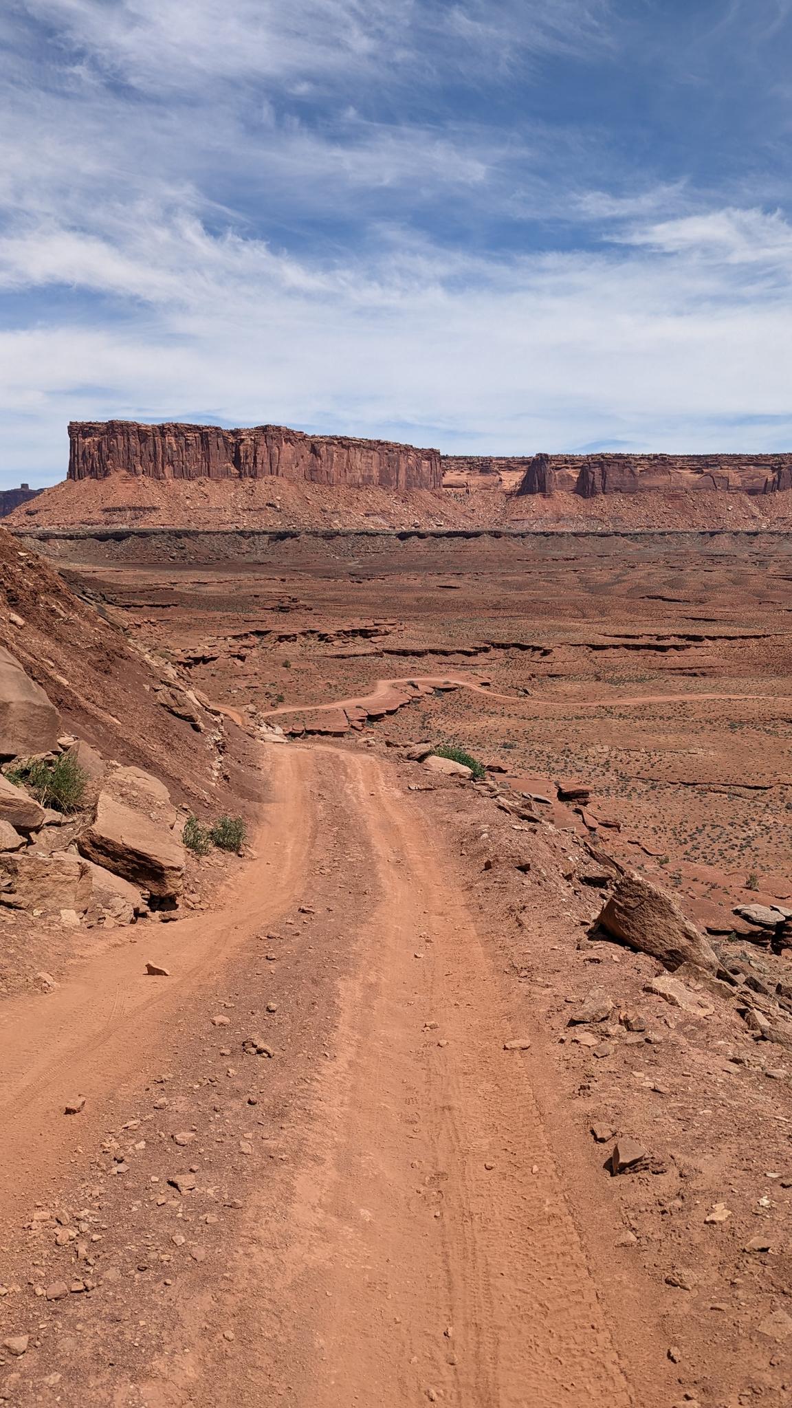 A wide dirt road leads to a large red rock mesa under a blue sky.