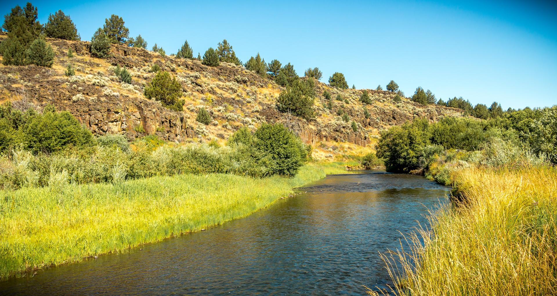 A calm river flows through a green valley with rocky, treed hills.