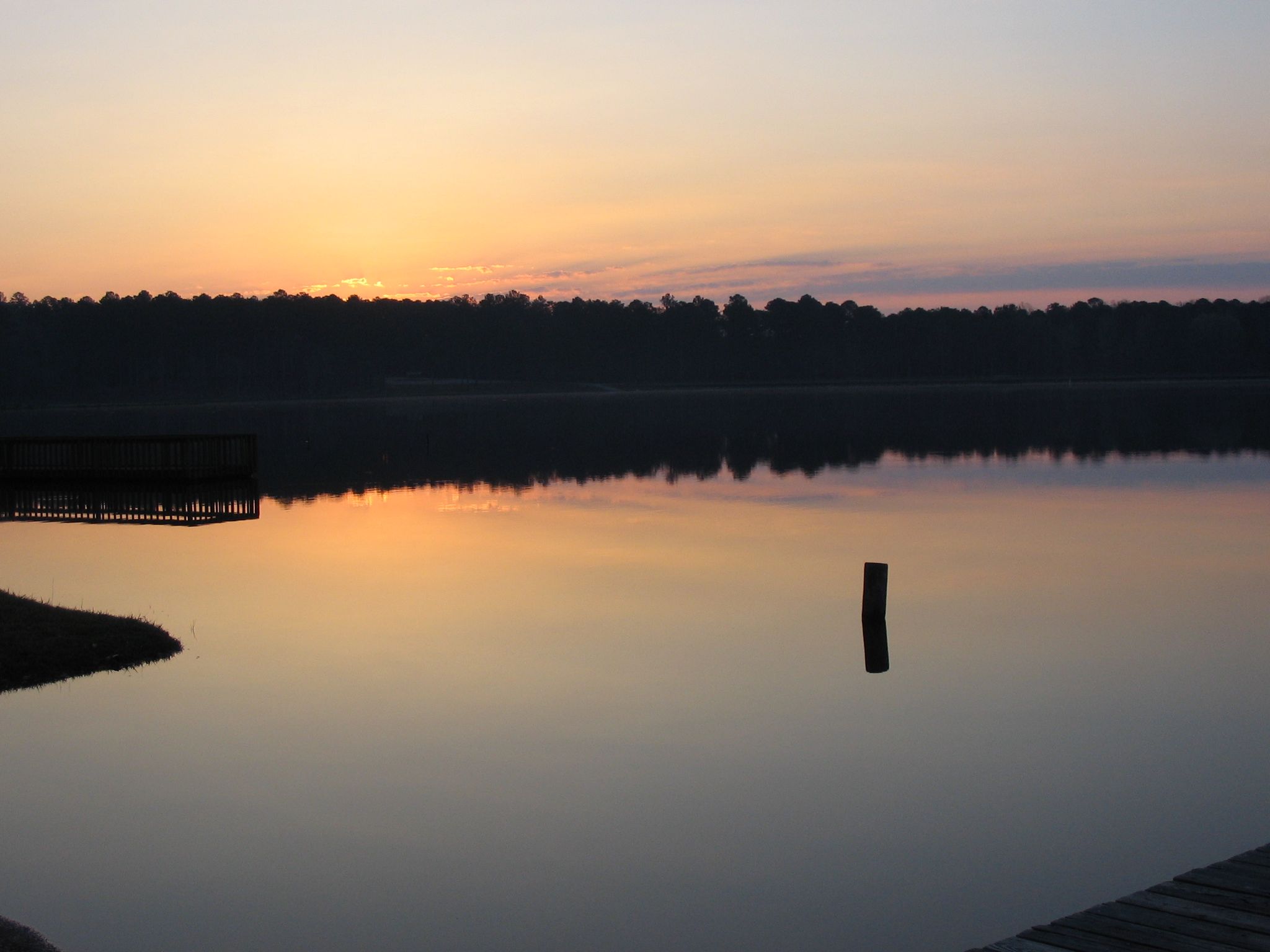 Calm lake reflecting a colorful sunrise sky and dark forest. - Noxubee Hills Outer Loop