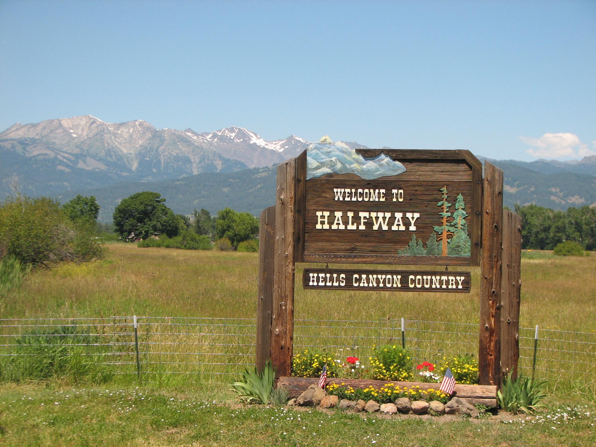 A wooden welcome sign stands in a sunny, grassy field with distant snow-capped mountains.