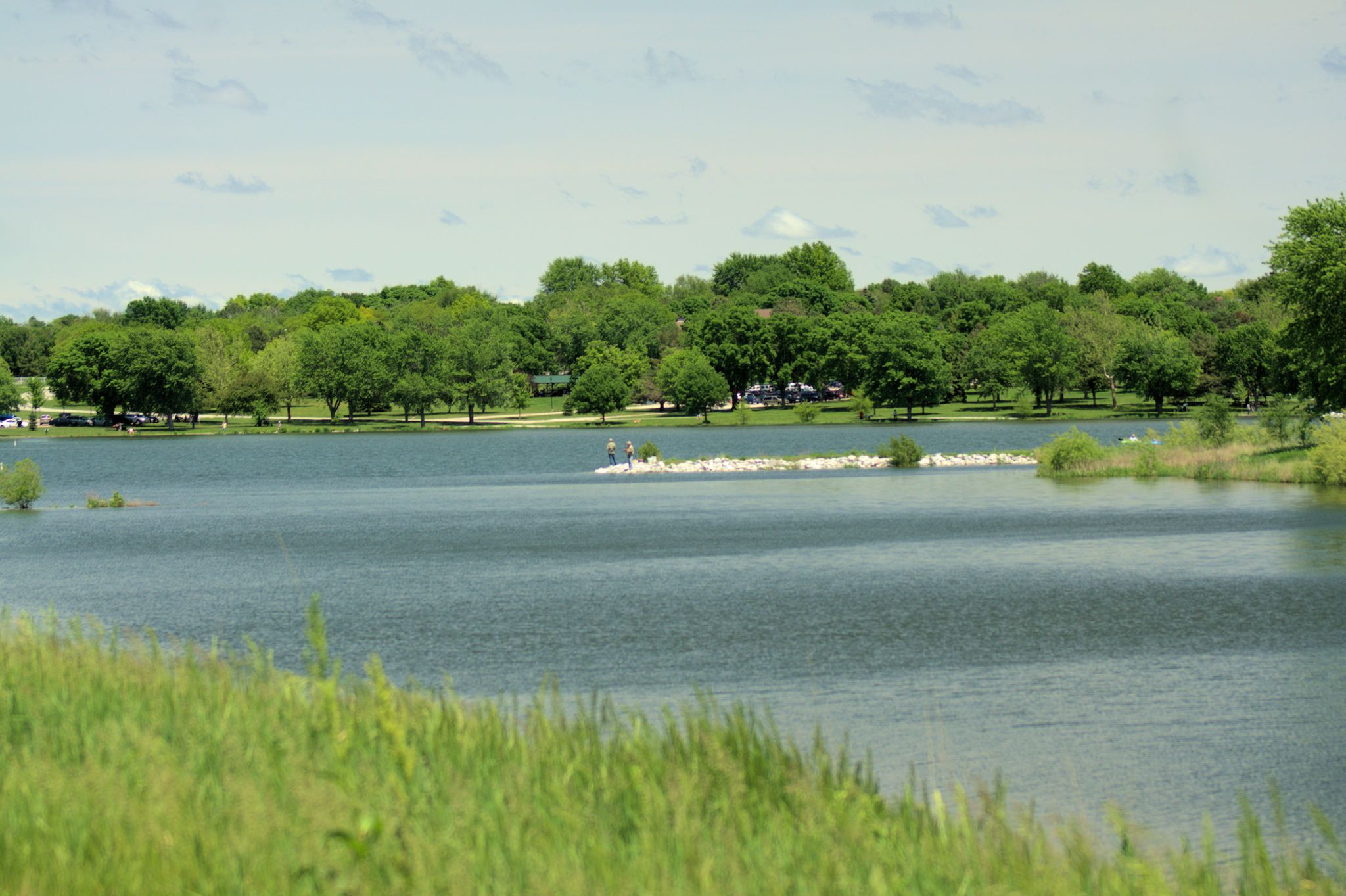 A calm lake with a small boat, surrounded by lush green trees under a bright sky. - Holmes Lake Park loop